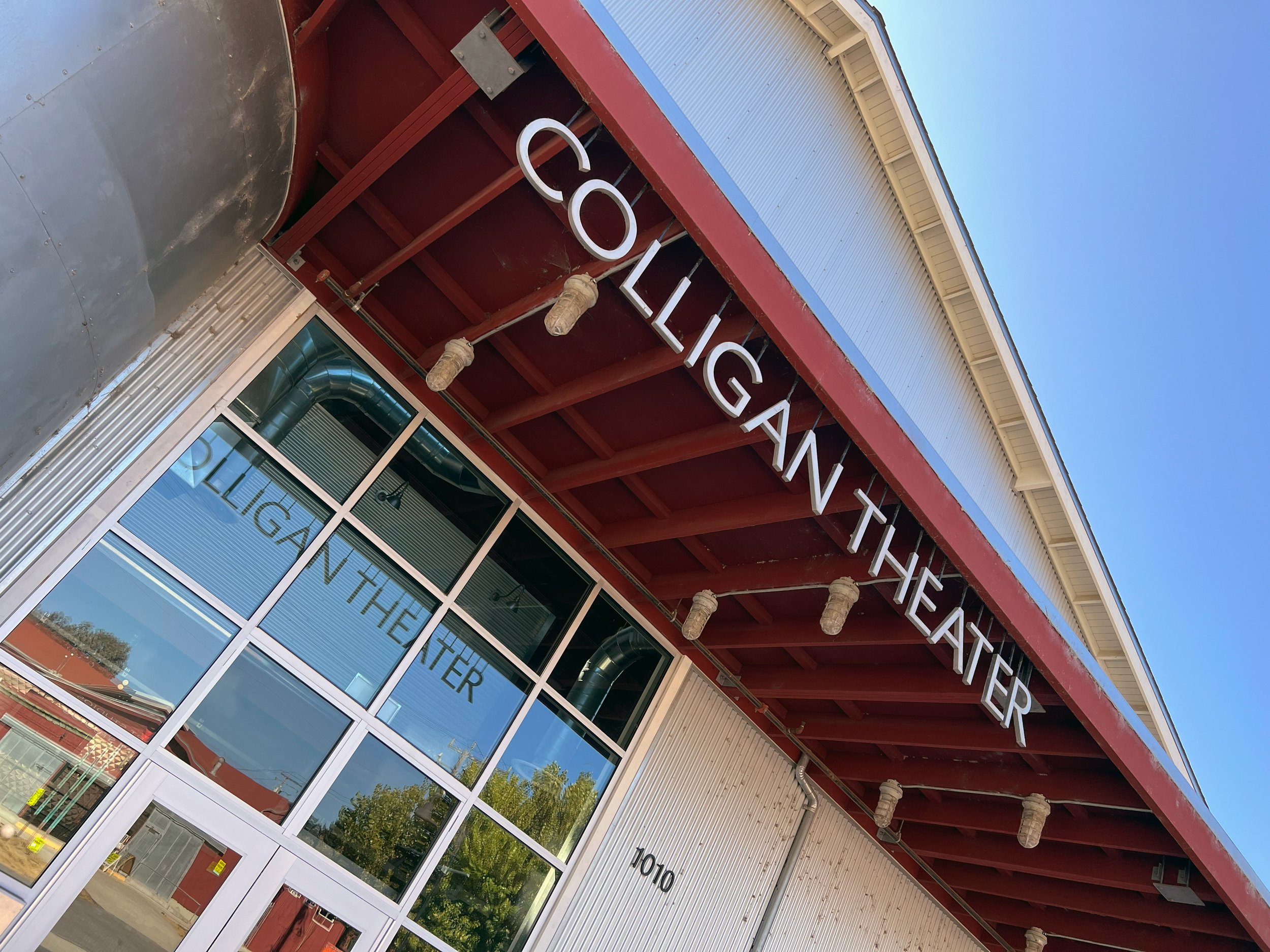 Exterior view of a building with large glass windows and a red and beige facade, featuring a sign that reads "COLOGIAN THEATER". The sky is clear and blue, and the reflection in the windows shows trees and neighboring structures.