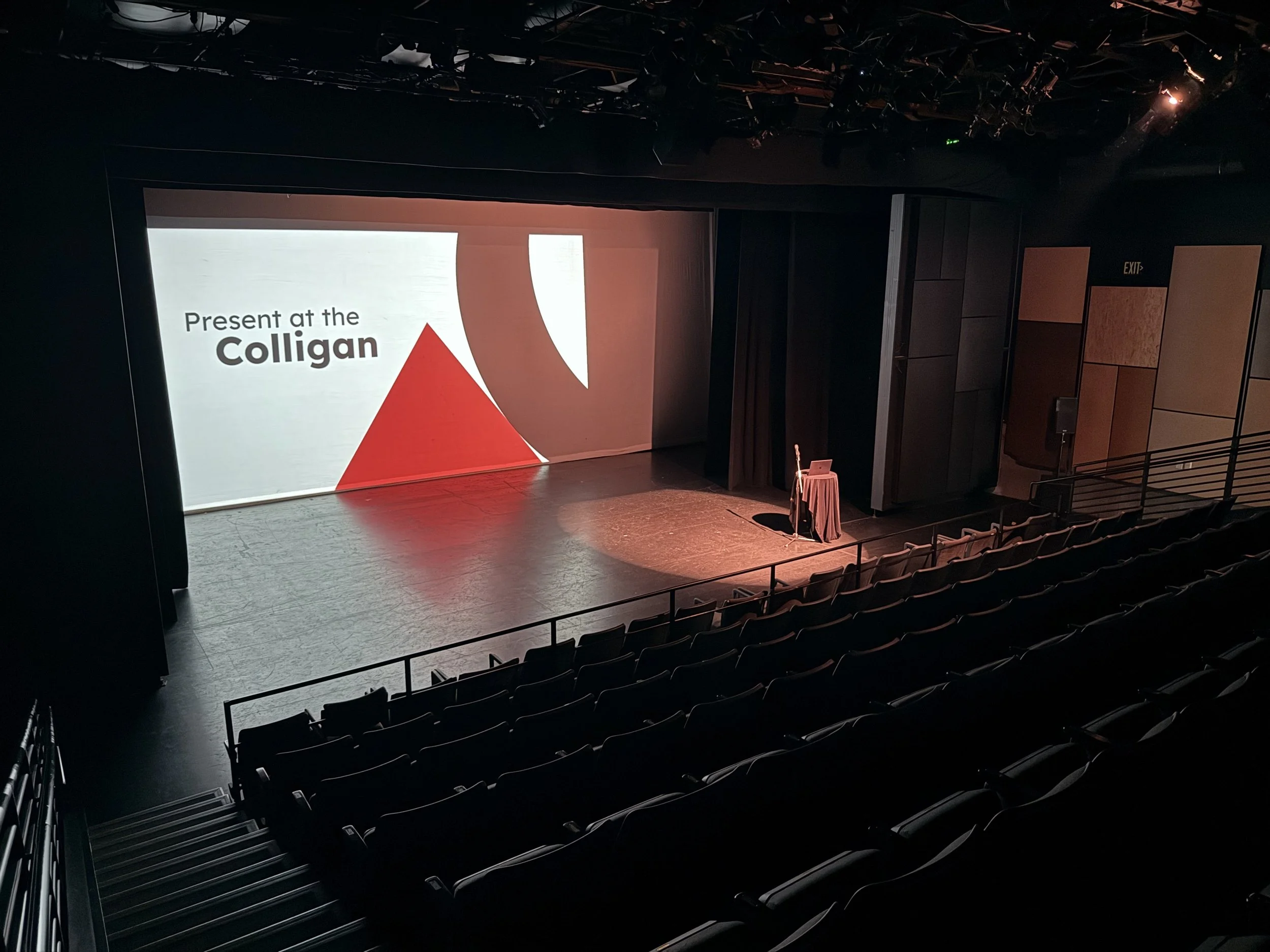 Empty theater auditorium with rows of black seats, a stage with a projector screen displaying the text 'Present at the Colligan' and a red triangle. There is a small table with a laptop and microphone on the stage.
