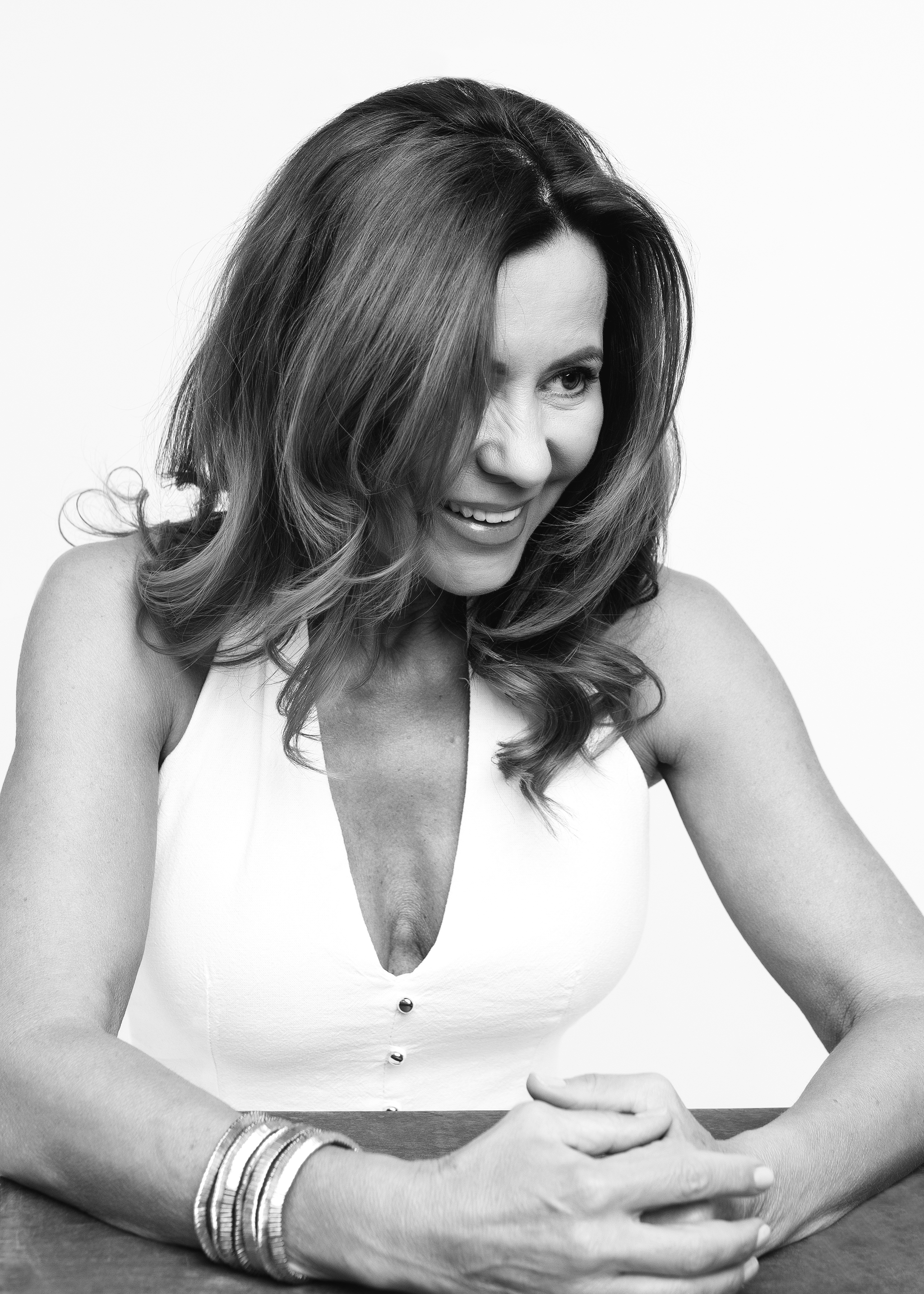 A woman smiling and looking to her left, with shoulder-length wavy hair, wearing a sleeveless white top with buttons, and multiple bracelets on her left wrist, sitting at a table.