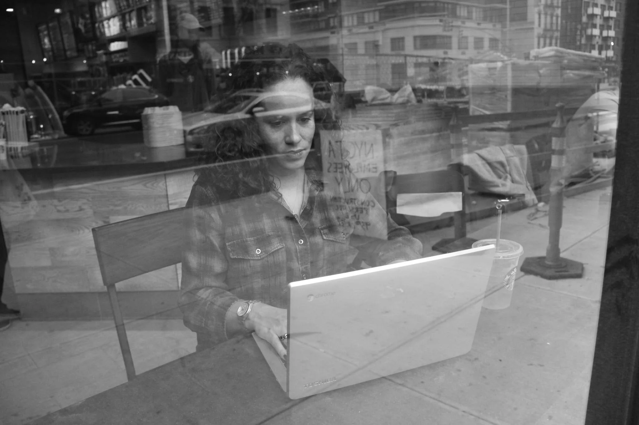 A woman with curly hair sitting at a table inside a coffee shop, working on a Chromebook laptop. Outside the window, cars and buildings are visible.