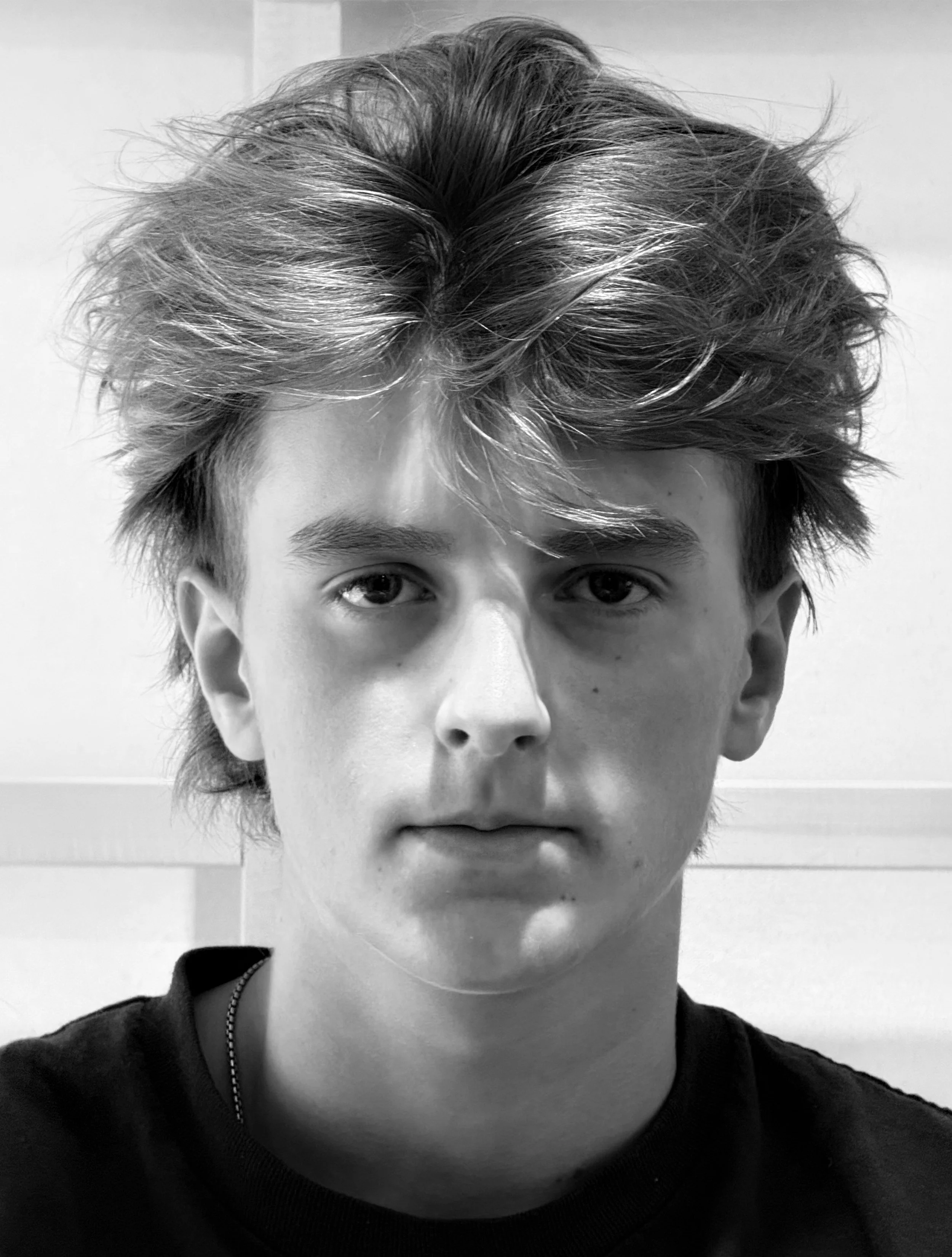 Black and white close-up portrait of a young man with voluminous, tousled hair, wearing a dark shirt, looking directly at the camera.