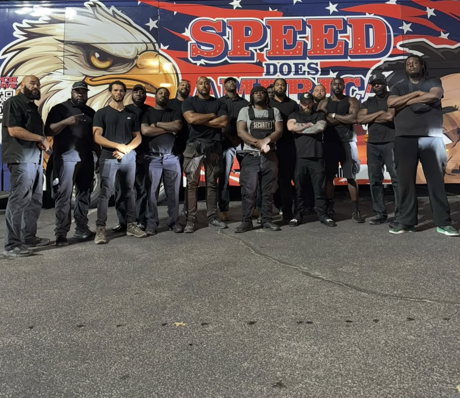 A group of approximately fifteen men standing together at night in front of a large colorful mural featuring an eagle and the words 'Speed Does Matter'. Most of the men are dressed in black clothing, with some wearing caps or hats, and one security personnel is wearing a vest labeled 'Security'.