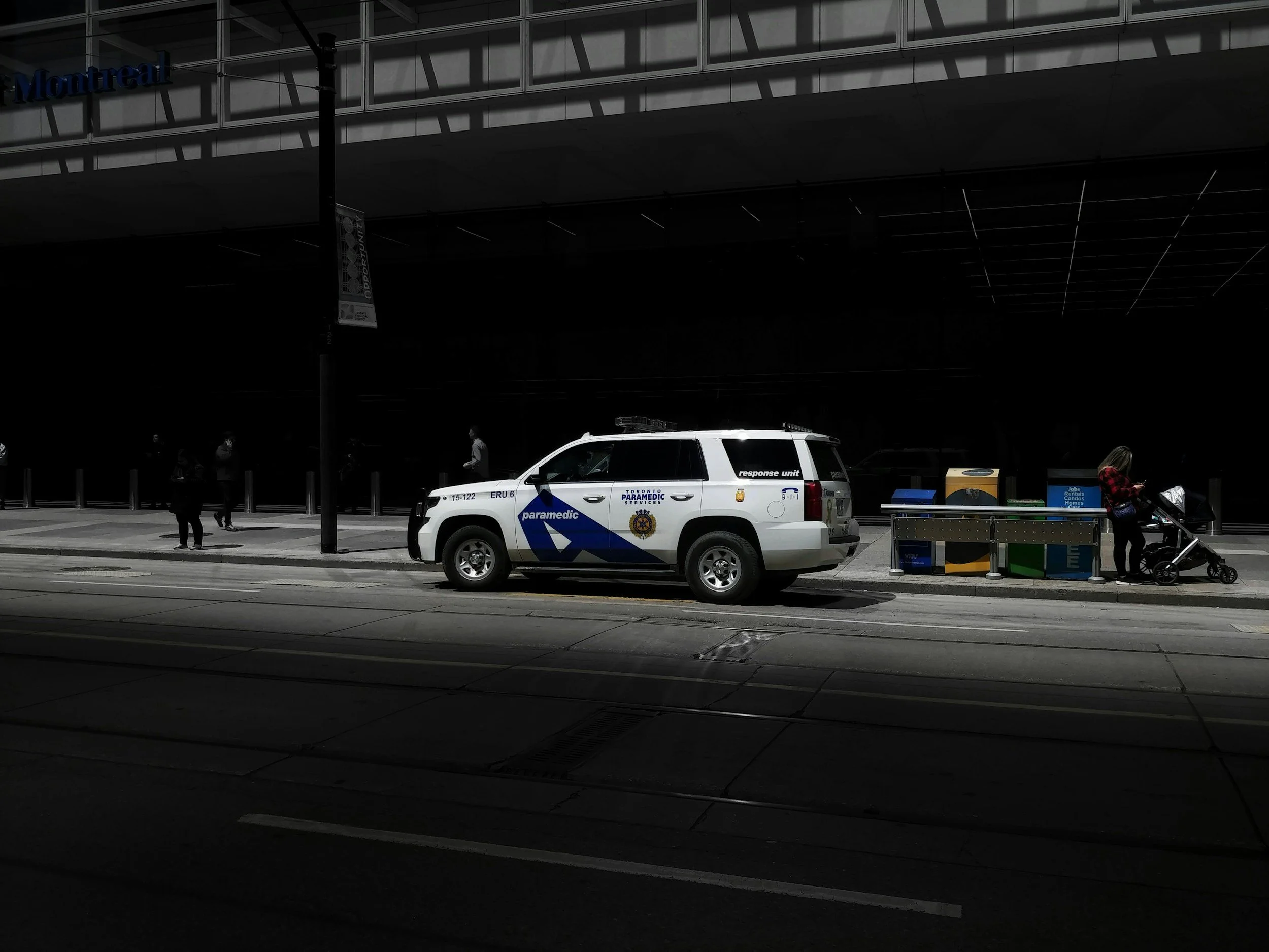 A white paramedic response vehicle parked on a city street near a sidewalk with a woman pushing a stroller and several pedestrians in the background.
