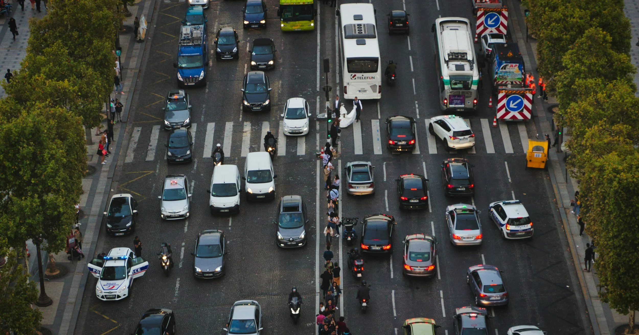 An aerial view of a busy city street with cars, buses, motorcycles, pedestrians, and street trees. There is a crosswalk in the center and roadwork signs on the right.