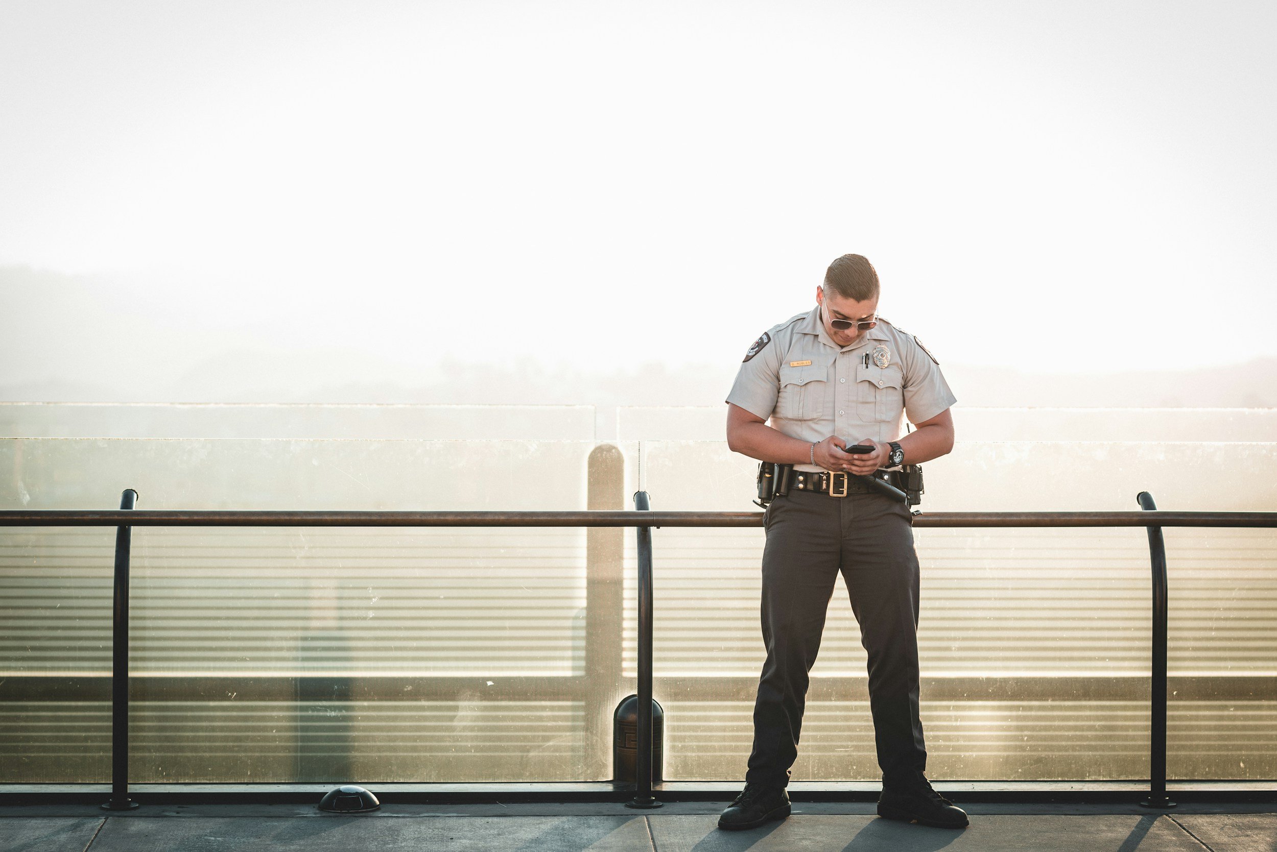 A police officer wearing sunglasses, a gray uniform, and black pants stands on a bridge, looking at his phone.