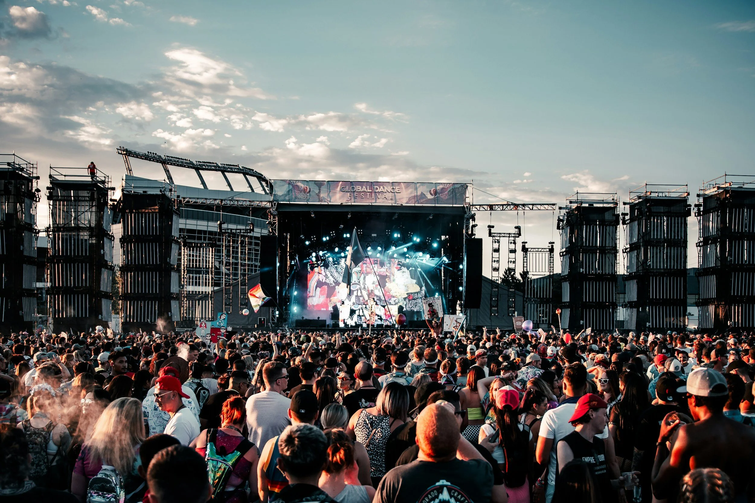 Crowd attending a music concert at an outdoor festival with a stage, performers, and colorful lighting under a partly cloudy sky.