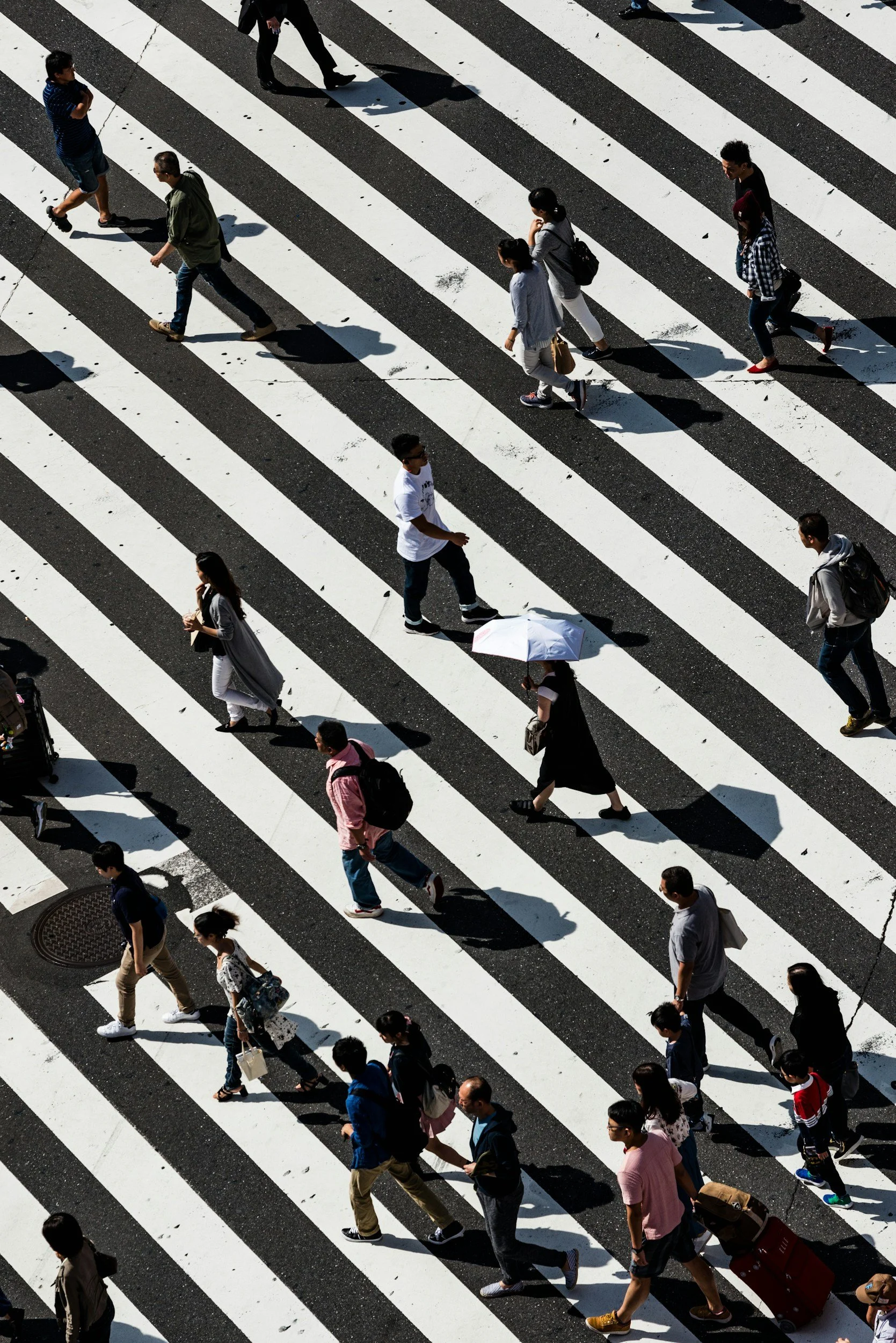 People crossing a black and white striped crosswalk on a city street, some carrying bags and using umbrellas.