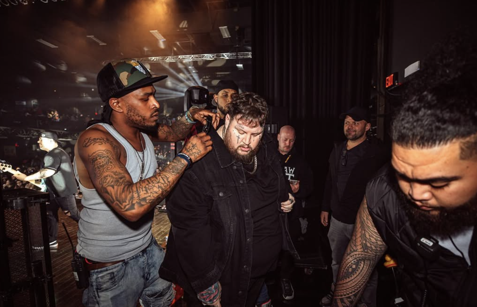 A man getting a haircut backstage at a concert with stage lights in the background.