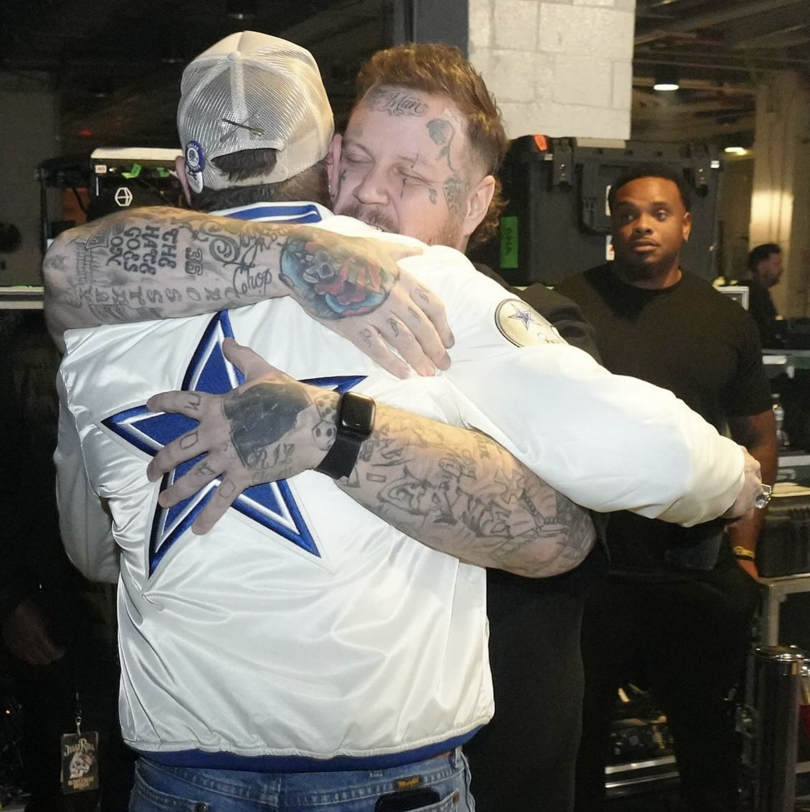 Two men hugging, one with numerous tattoos and the other wearing a Dallas Cowboys jacket and baseball cap, in an indoor setting with a person in the background observing.