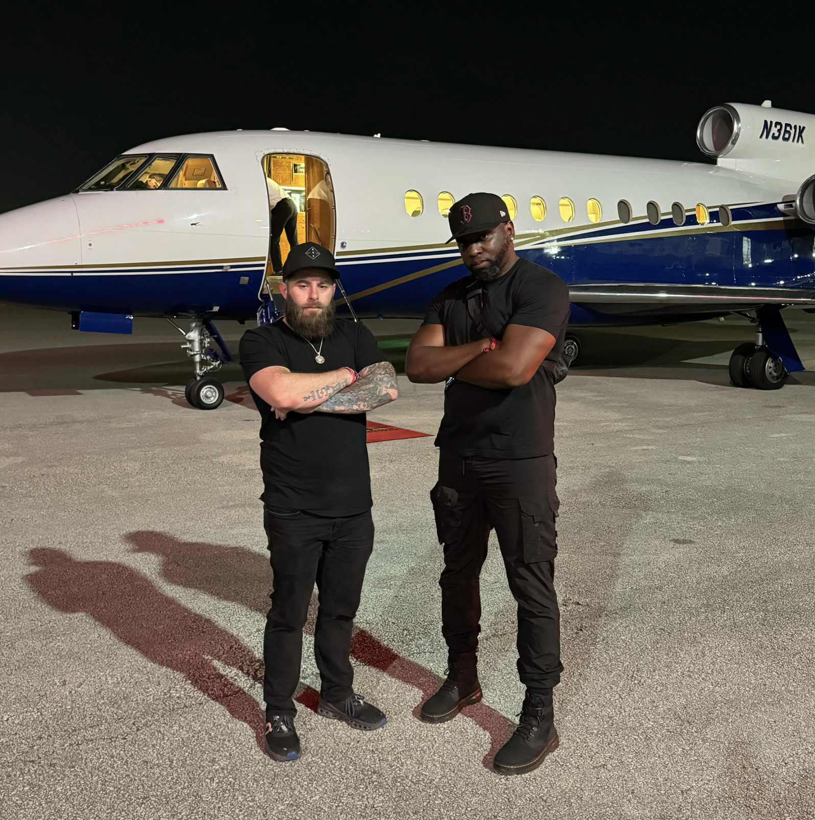 Two men standing in front of a private jet on the tarmac at night, with the jet's open door behind them. Both men are wearing black clothing and caps, and have arms crossed.