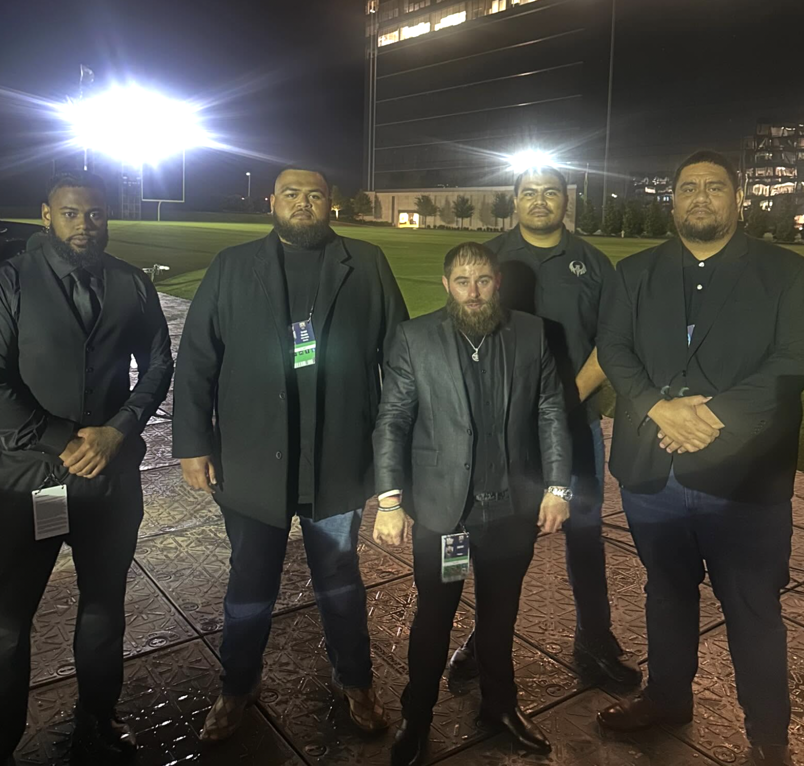 Group of five men in formal attire standing outdoors at night on a patterned sidewalk with bright stadium lights and a large building in the background.