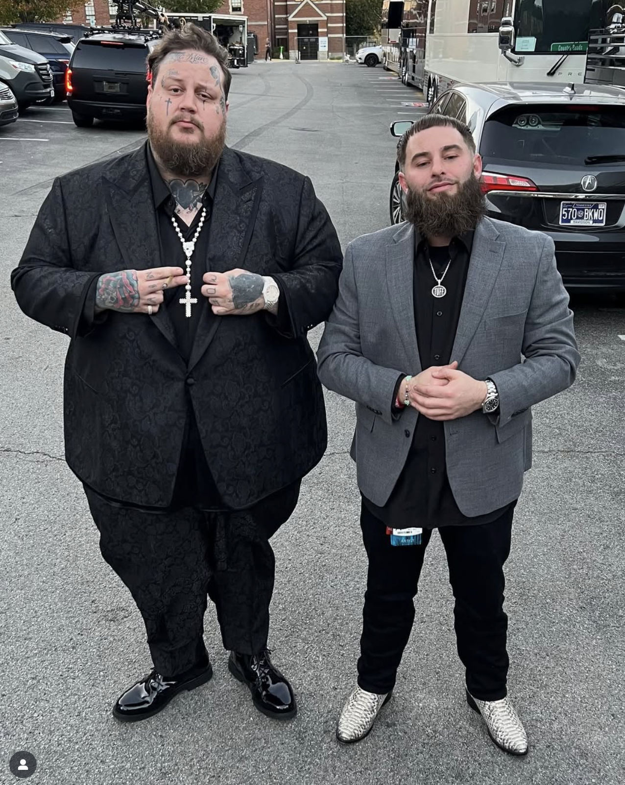 Two men standing in a parking lot, dressed in formal suits with tattoos. The man on the left wears a black patterned suit, shiny black shoes, and has tattoos on his face, hands, and neck. The man on the right wears a gray suit jacket, black shirt, an