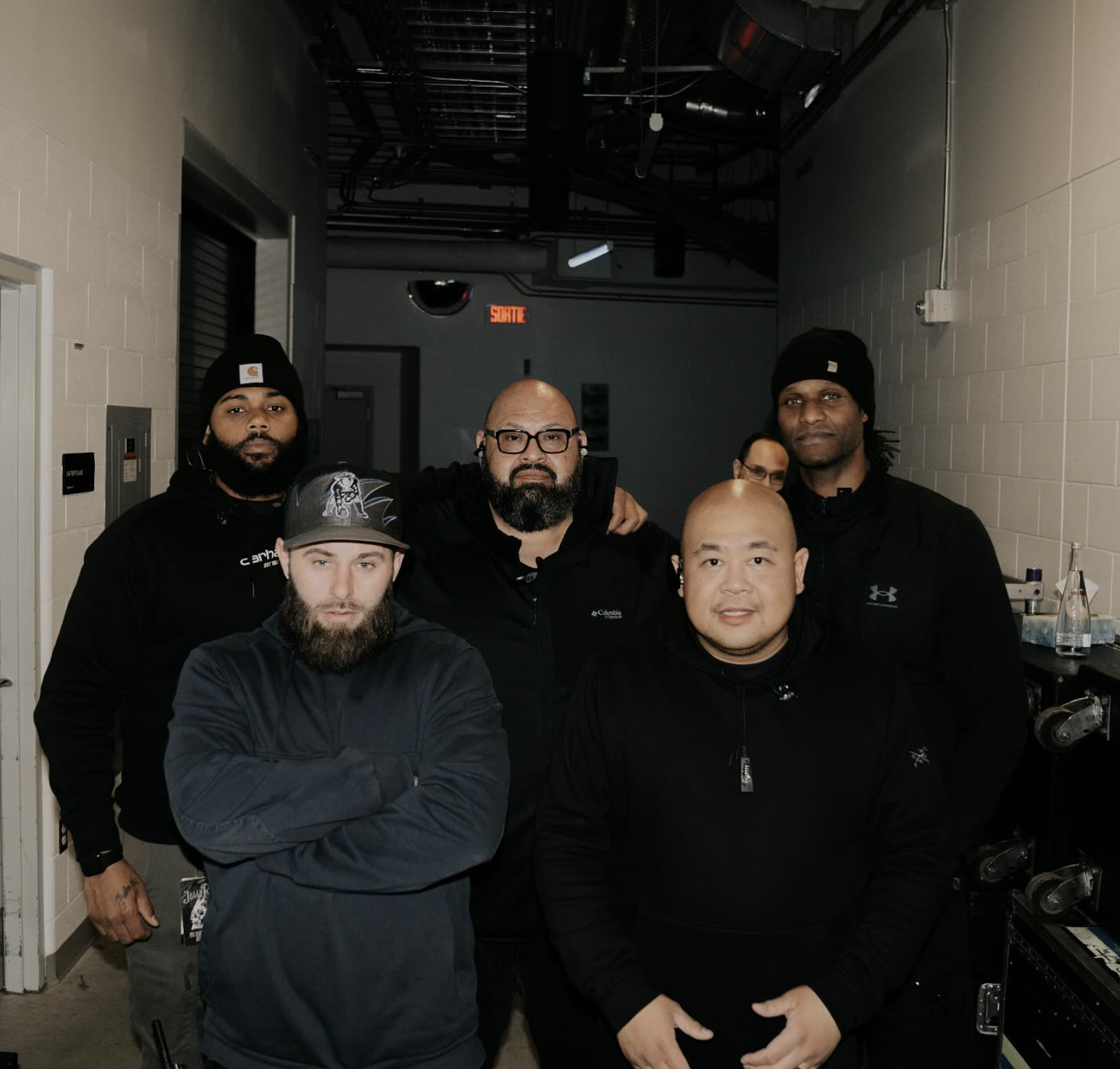 Group of five men standing together in a hallway, dressed in dark casual clothing, some wearing beanies and hoodies. They pose for the photo with serious expressions.