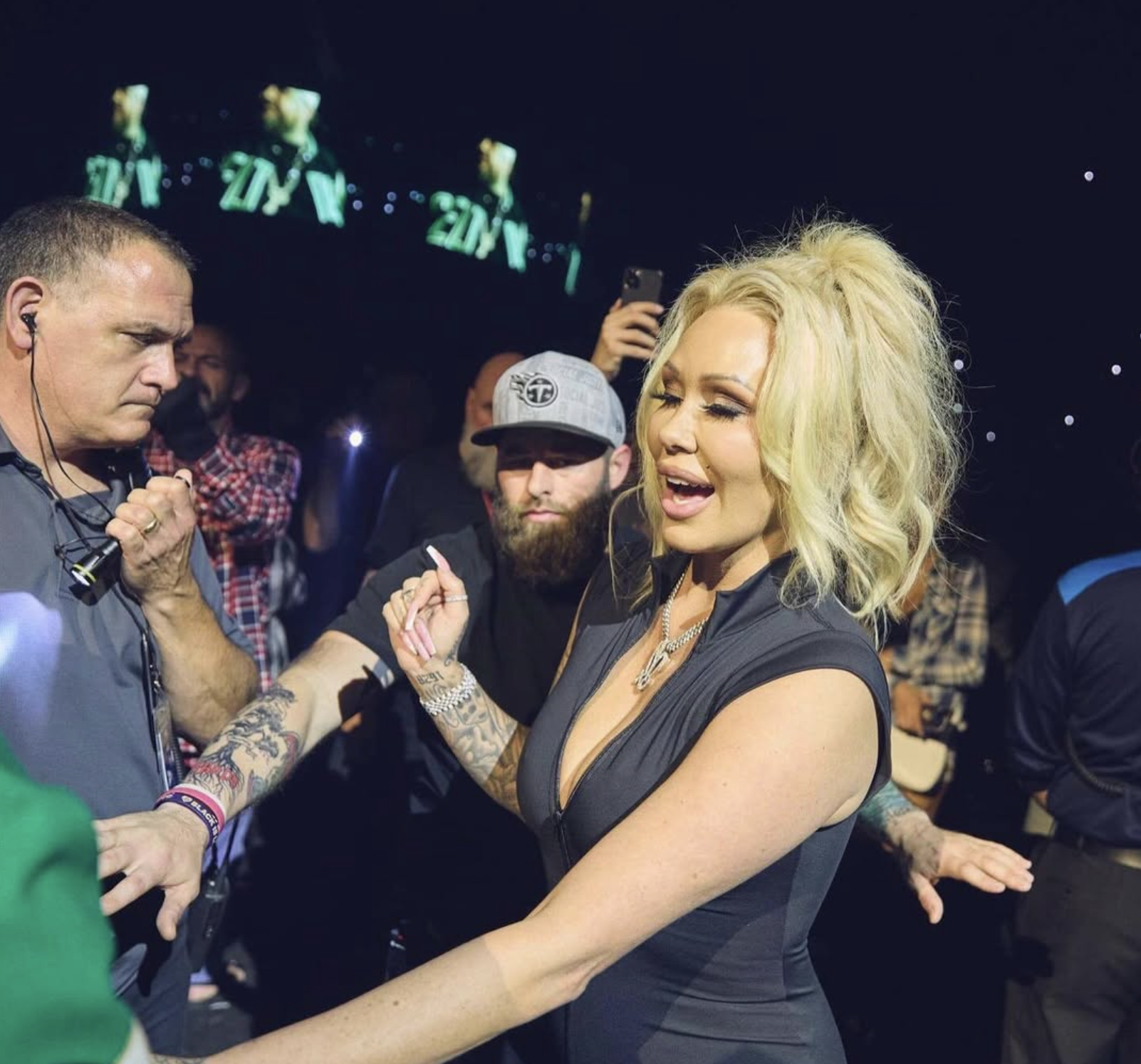 A woman with blonde, curly hair singing at a concert, surrounded by people, including a tattooed man and others in the background.