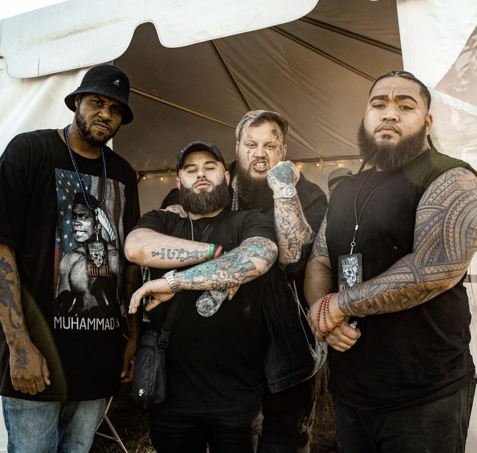 Four men with tattoos posing together in a tent at an event, with one wearing a Muhammad T-shirt and others with beards and distinctive hairstyles.