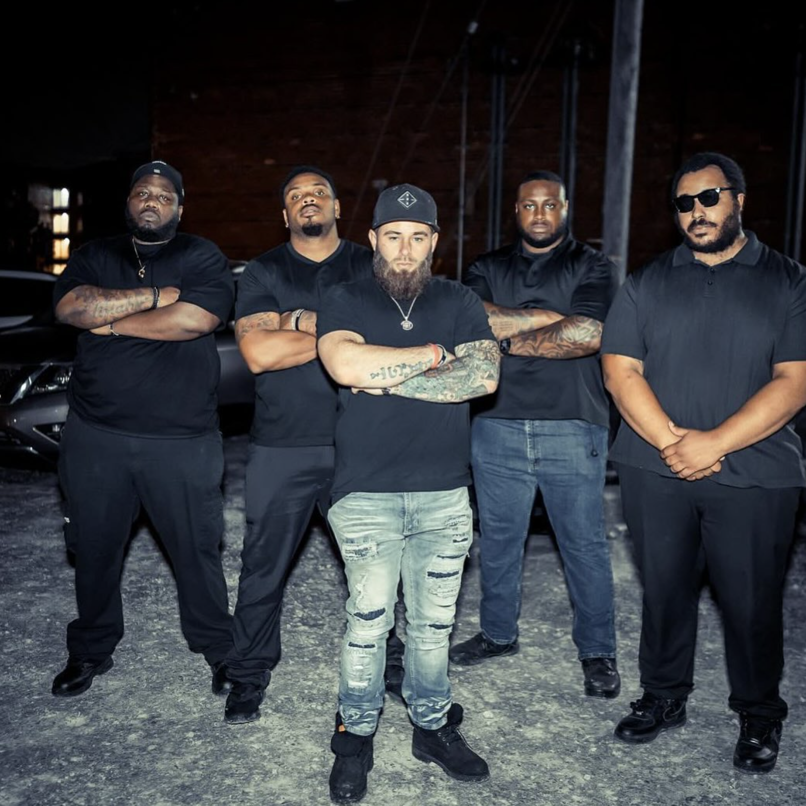 Five men standing outdoors at night, wearing black shirts and posing with crossed arms in front of dark background and cars.