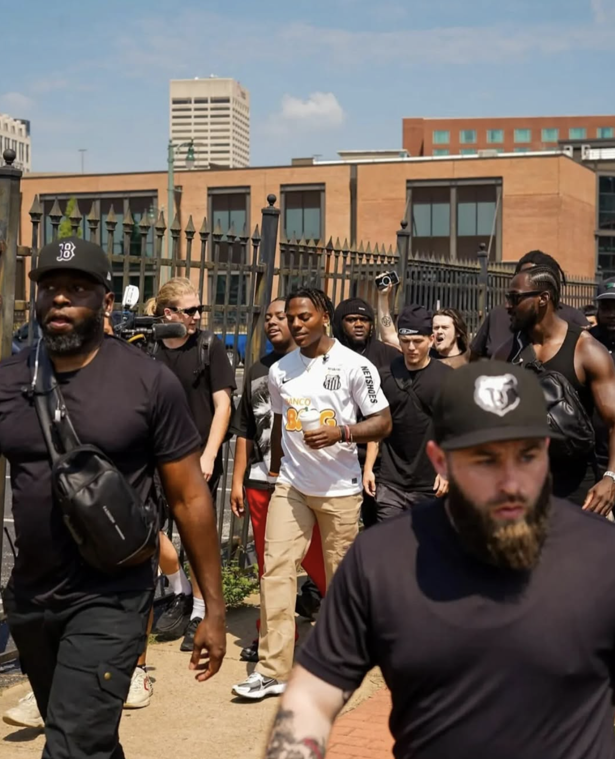 A group of people walking outdoors near a black metal fence with brick and modern buildings in the background, some wearing black shirts, and one man in a white soccer jersey holding a cup.