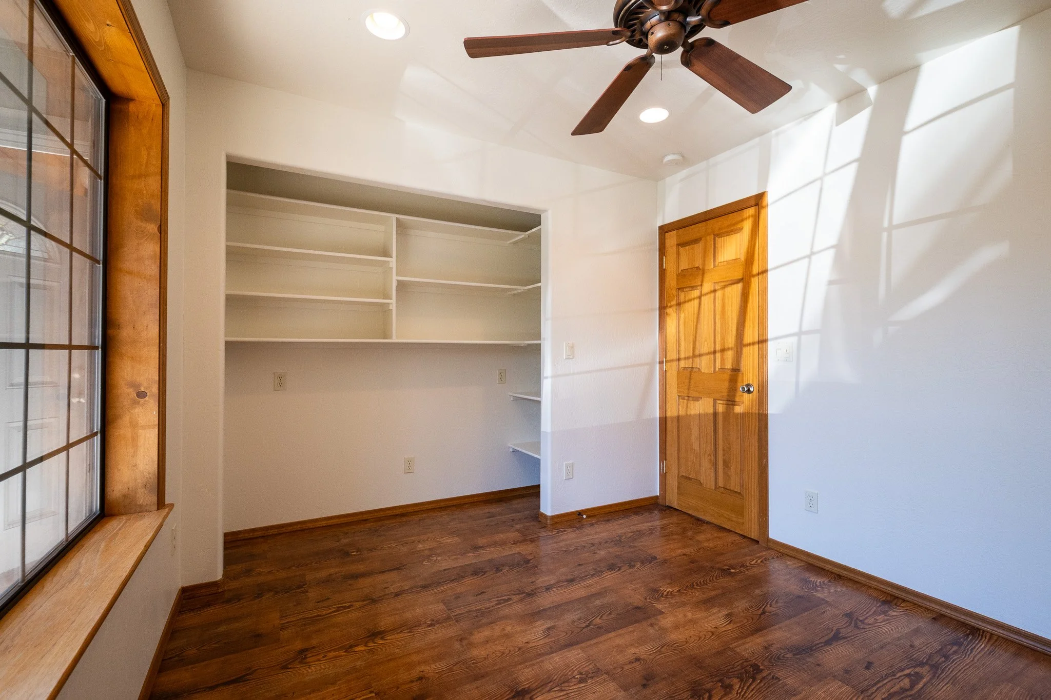 Empty room with hardwood floor, white walls, a wooden door, a ceiling fan, built-in shelves, and a window with a wooden frame.