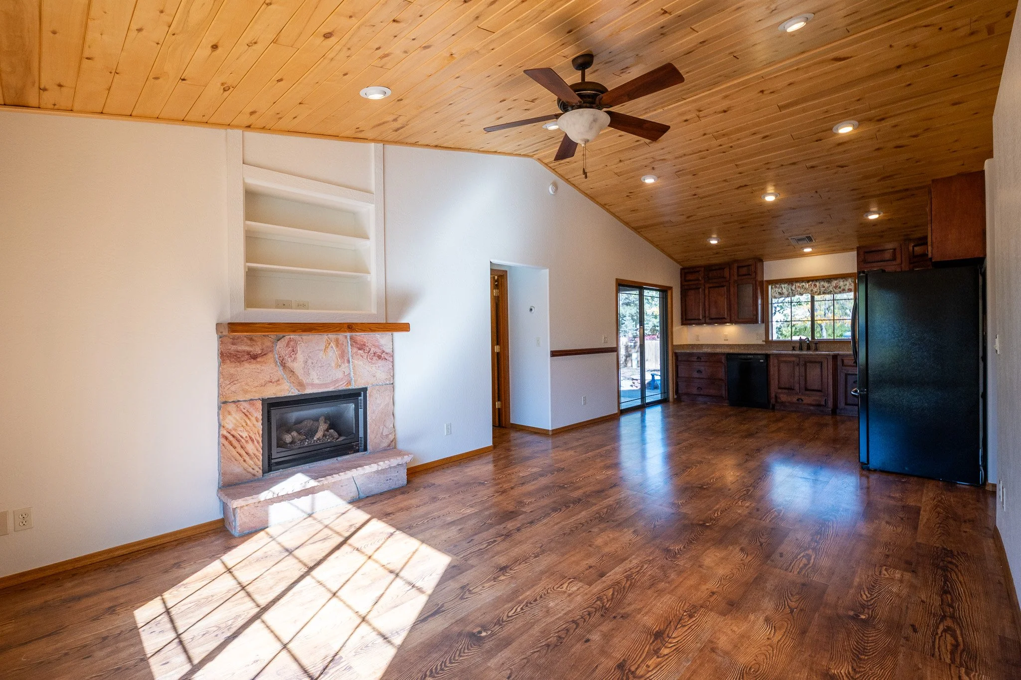 Interior view of a living room with a vaulted wooden ceiling, a ceiling fan, hardwood floors, a fireplace with a stone surround, and kitchen cabinets in the background.