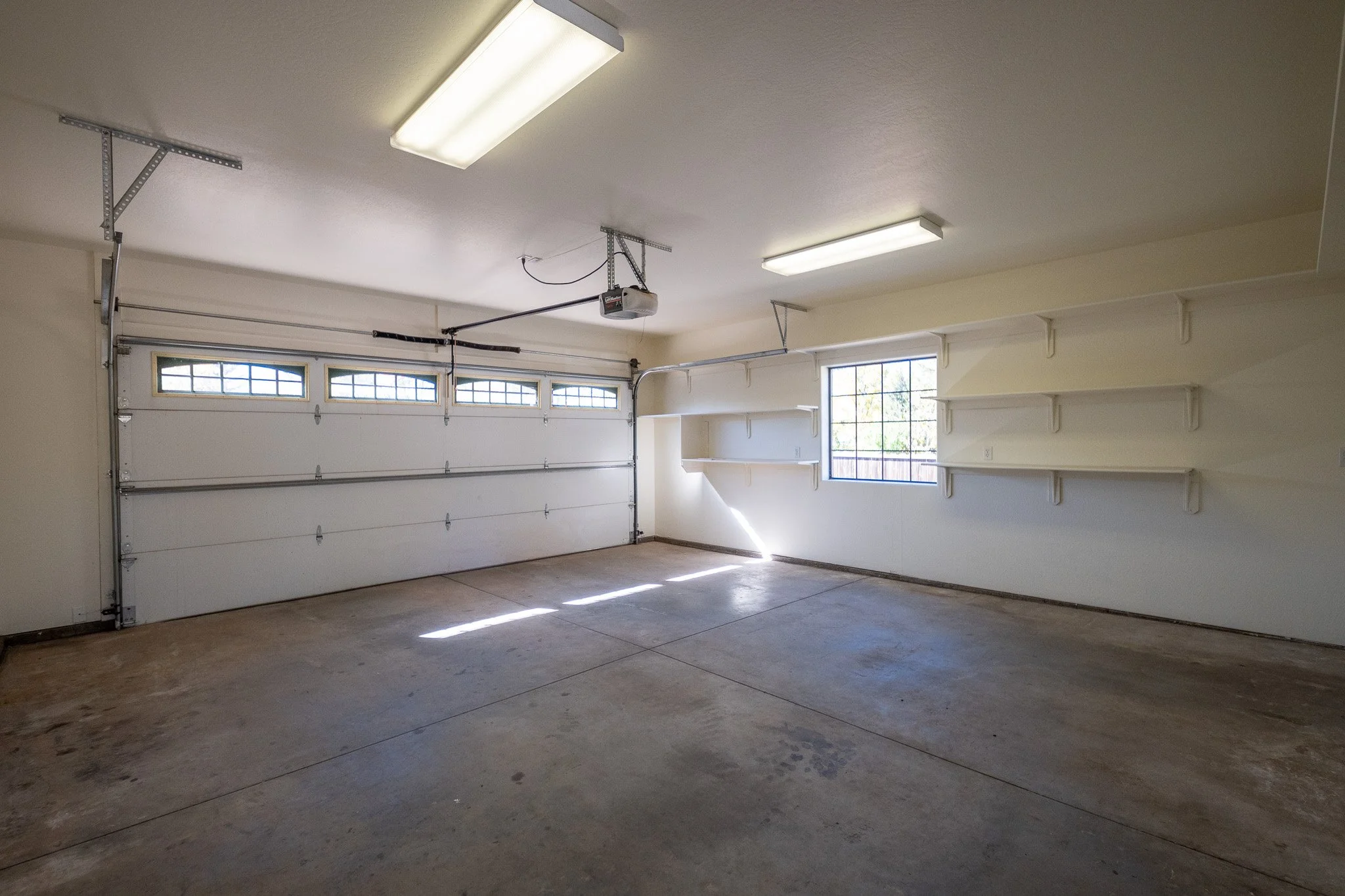 Empty garage with concrete floors, white walls, large white sectional garage door with windows, overhead lighting, and white shelving along the wall.