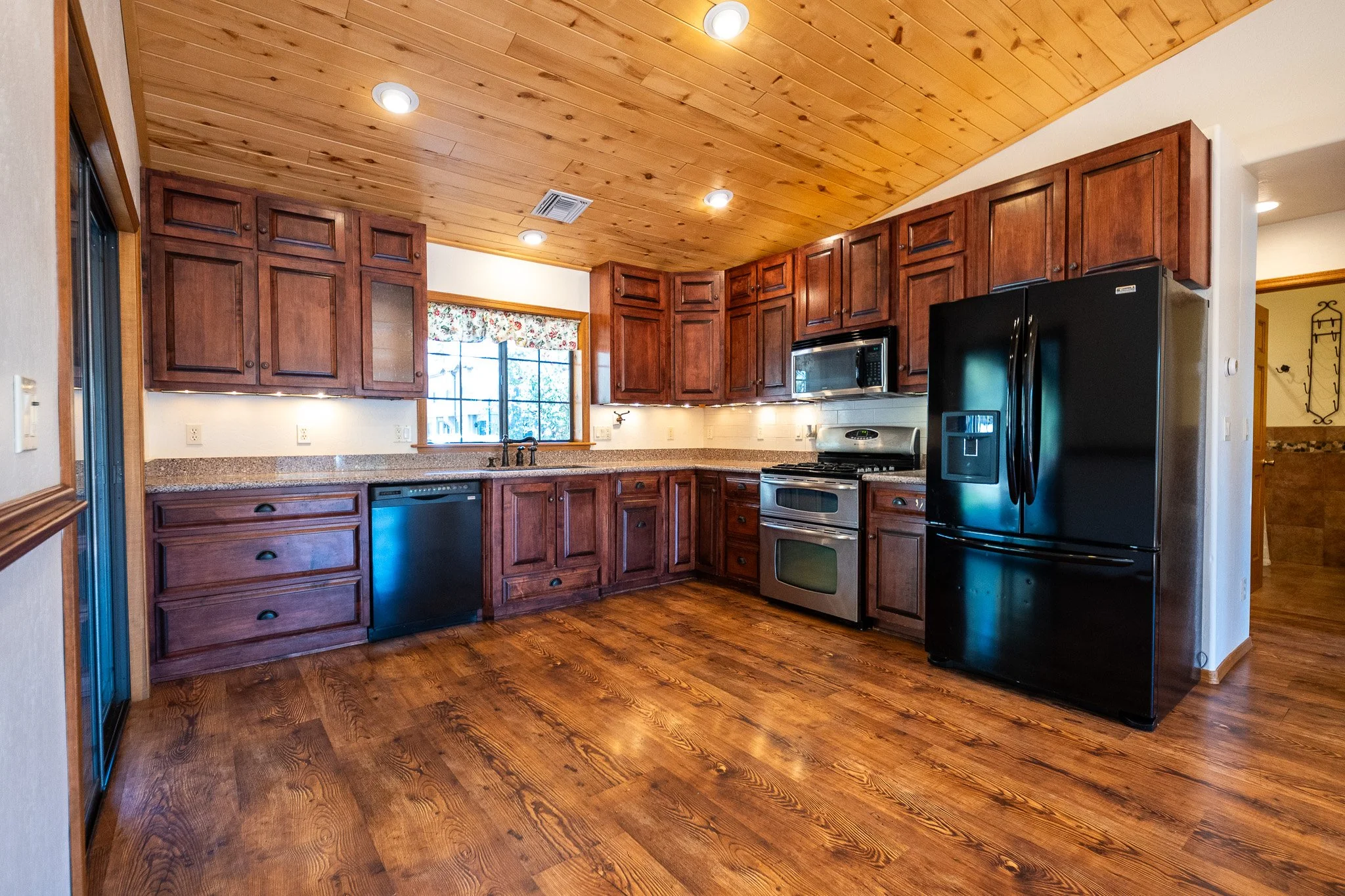 Kitchen with wooden cabinets, granite countertops, stainless steel appliances, black refrigerator, gas stove, microwave, wood flooring, wooden ceiling with recessed lighting, window with floral curtain.
