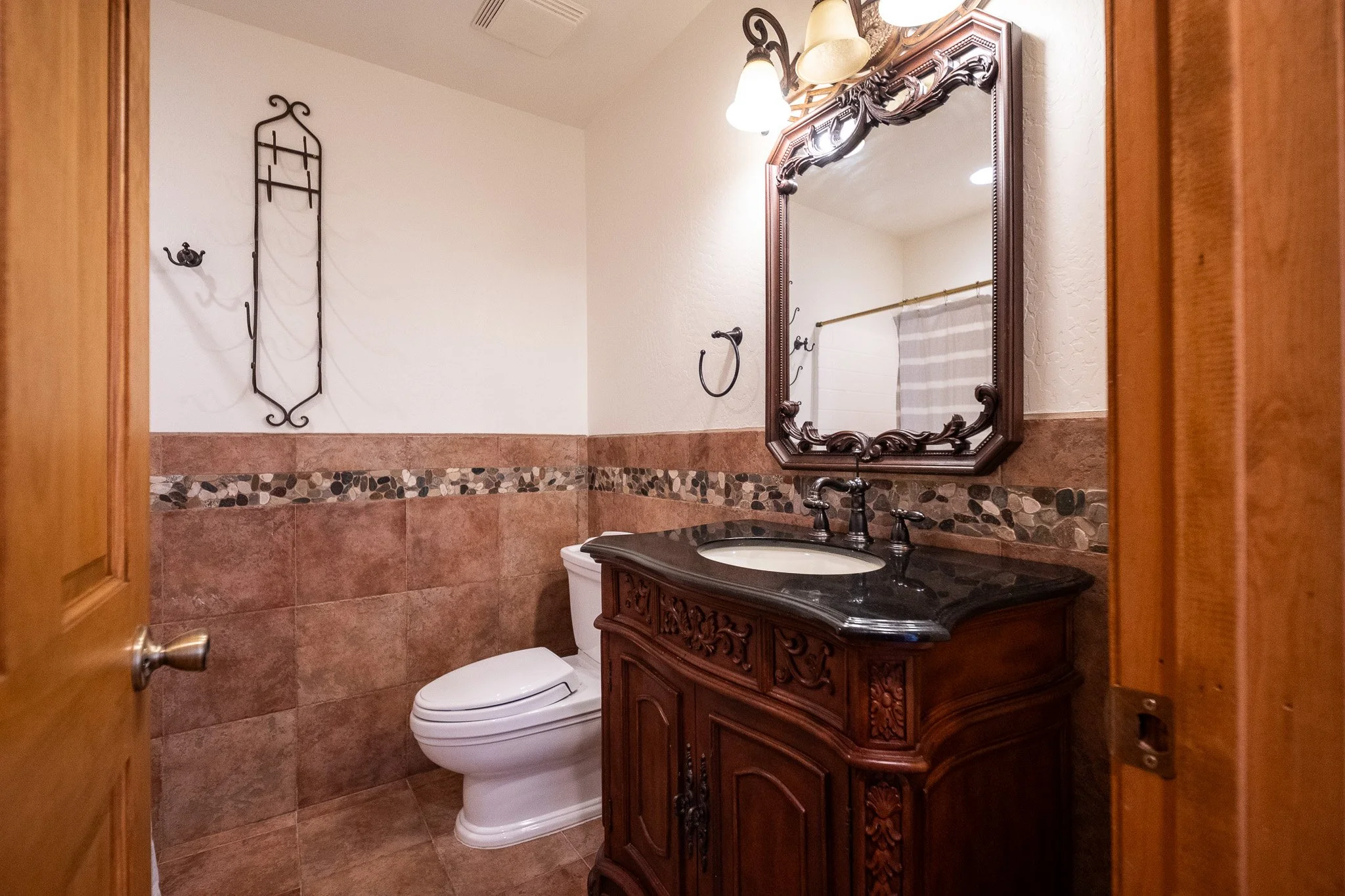 A bathroom with a decorative wooden vanity with a black countertop and a white sink, a large ornate mirror, vintage-style lighting, a toilet, a wall-mounted decorative iron coat rack, and a white and gray striped shower curtain.