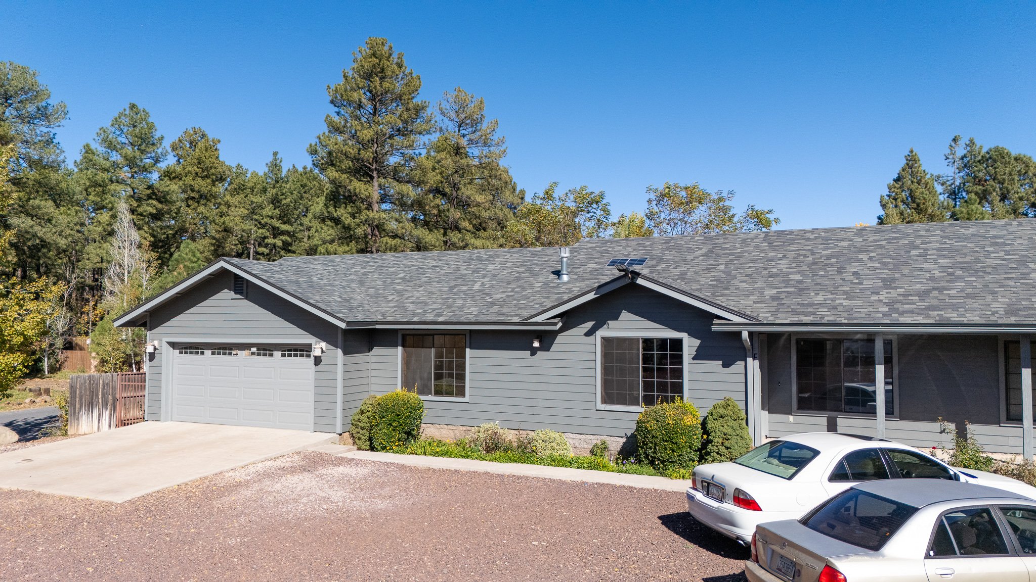 Front view of a gray house with a two-car garage, a driveway, and two parked cars, with a background of tall pine trees and a blue sky.