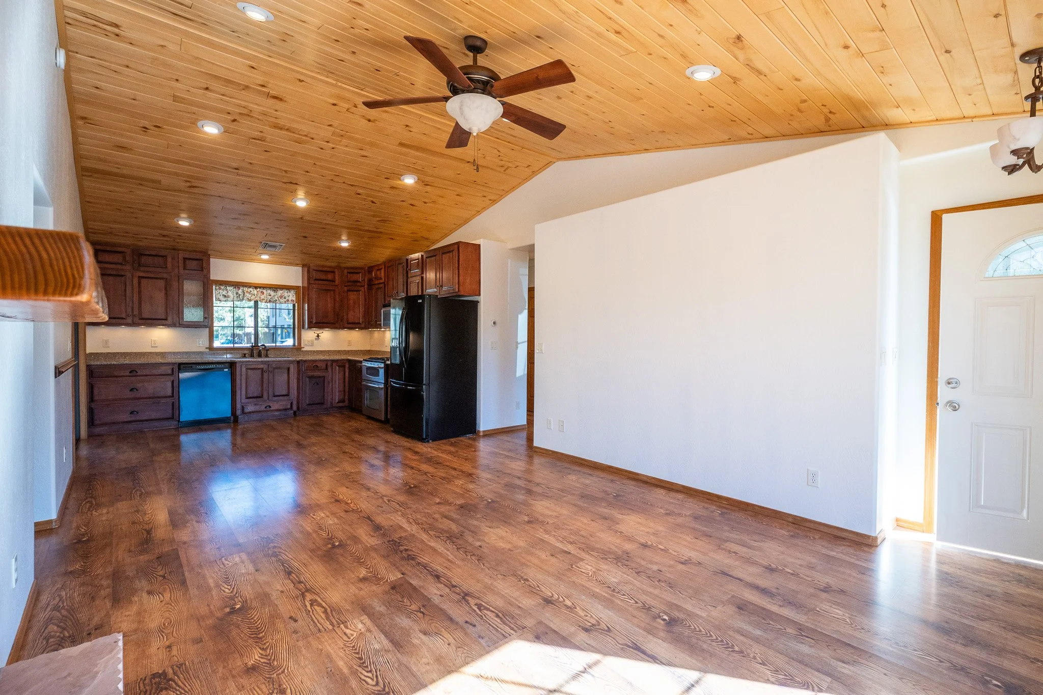 A room with a wooden ceiling, hardwood flooring, and a kitchen with dark wood cabinets, a black refrigerator, and a window with a floral valance. Recessed and hanging lights are visible.