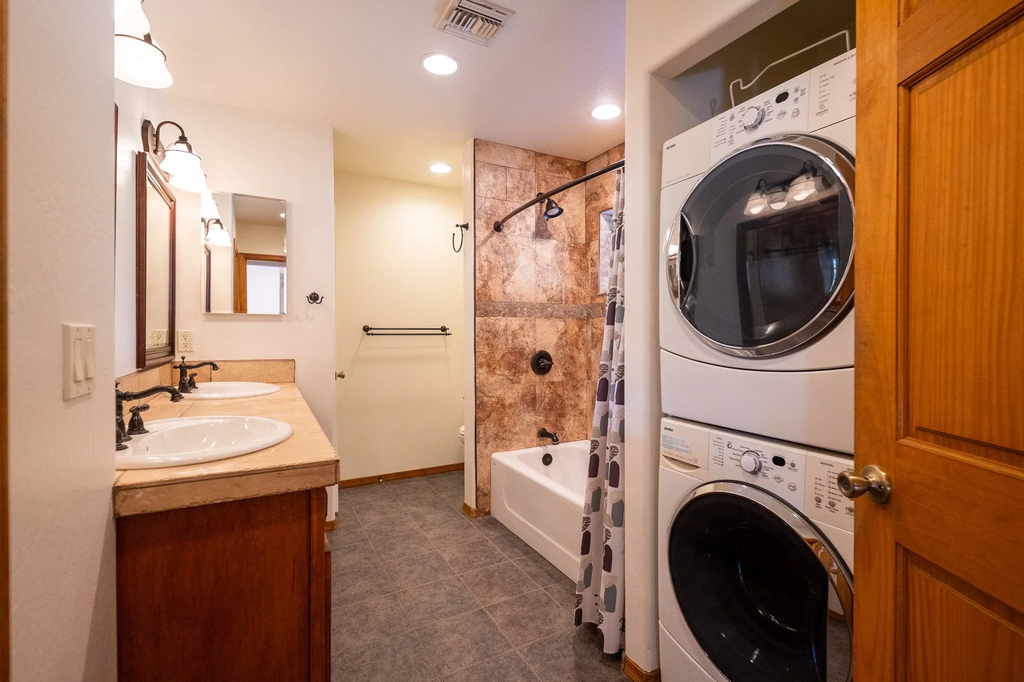 A bathroom with a washer and dryer stacked on top of each other, a bathtub with a shower curtain, double sinks with mirrors and lights, and a wall with brown tiles.