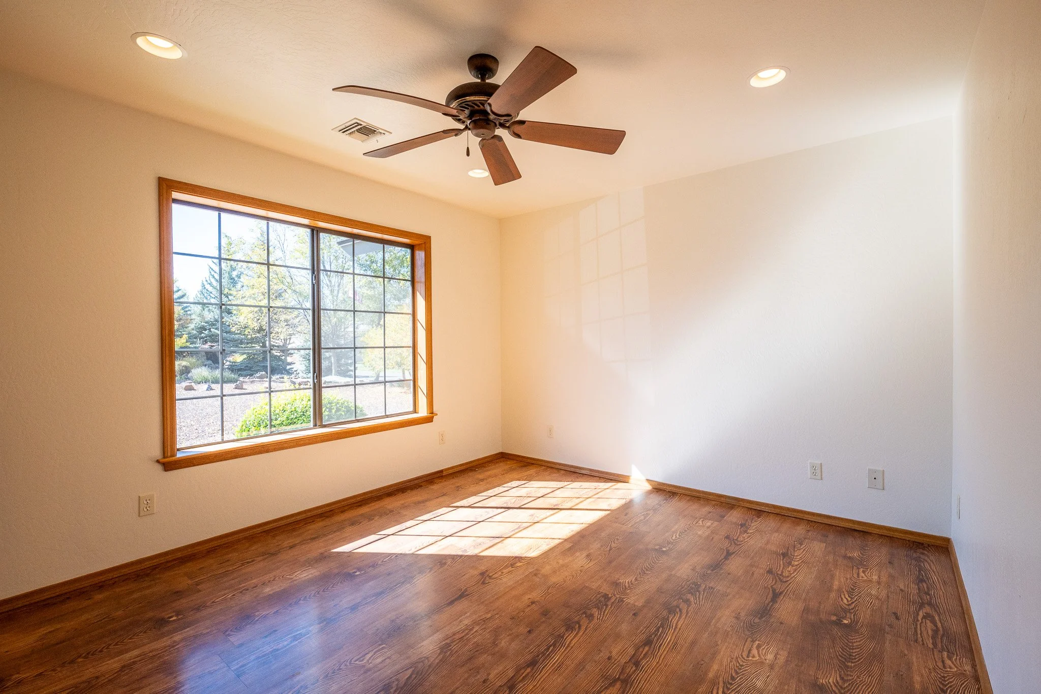 Empty room with hardwood floors, a large window with wooden trim, ceiling fan, and recessed lighting.
