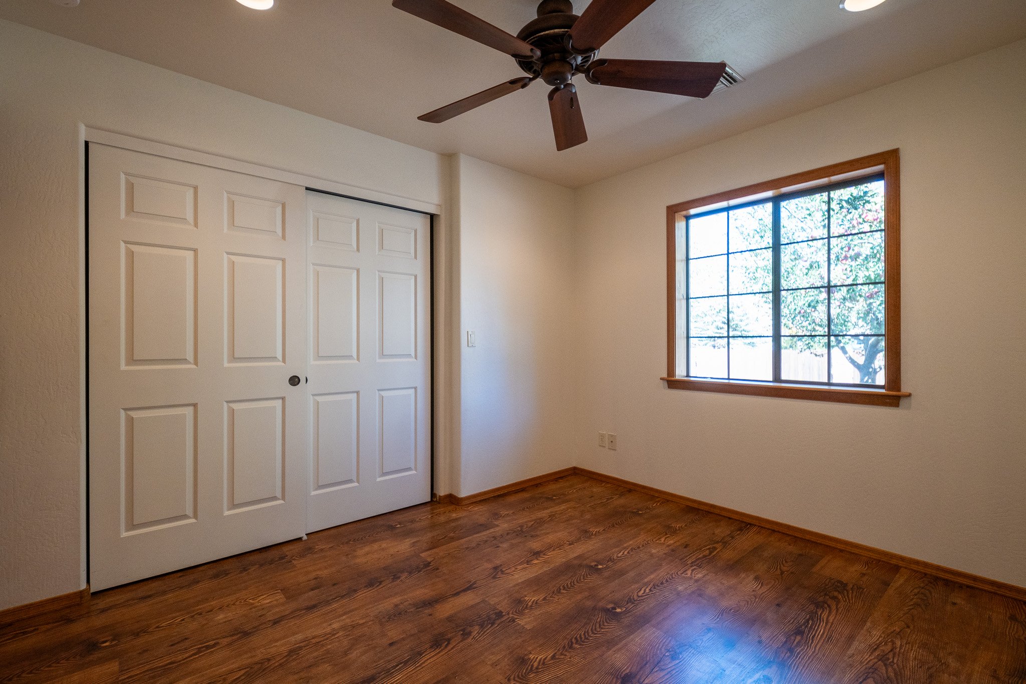 Room with a closet door, a window showing trees outside, a ceiling fan, and hardwood floors.