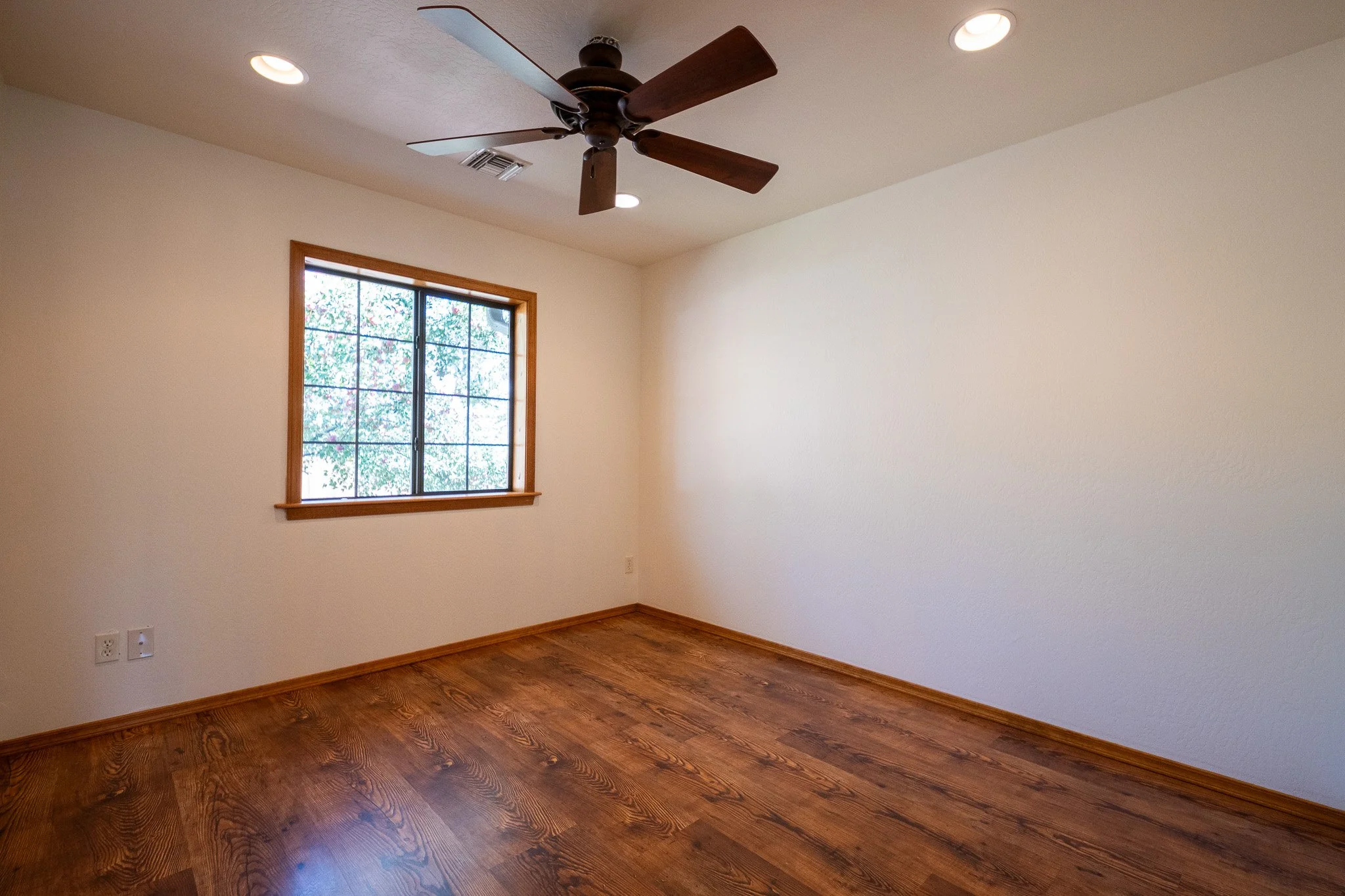 An empty room with beige walls, hardwood floors, a window with wooden trim, a ceiling fan with wooden blades, and recessed lighting.