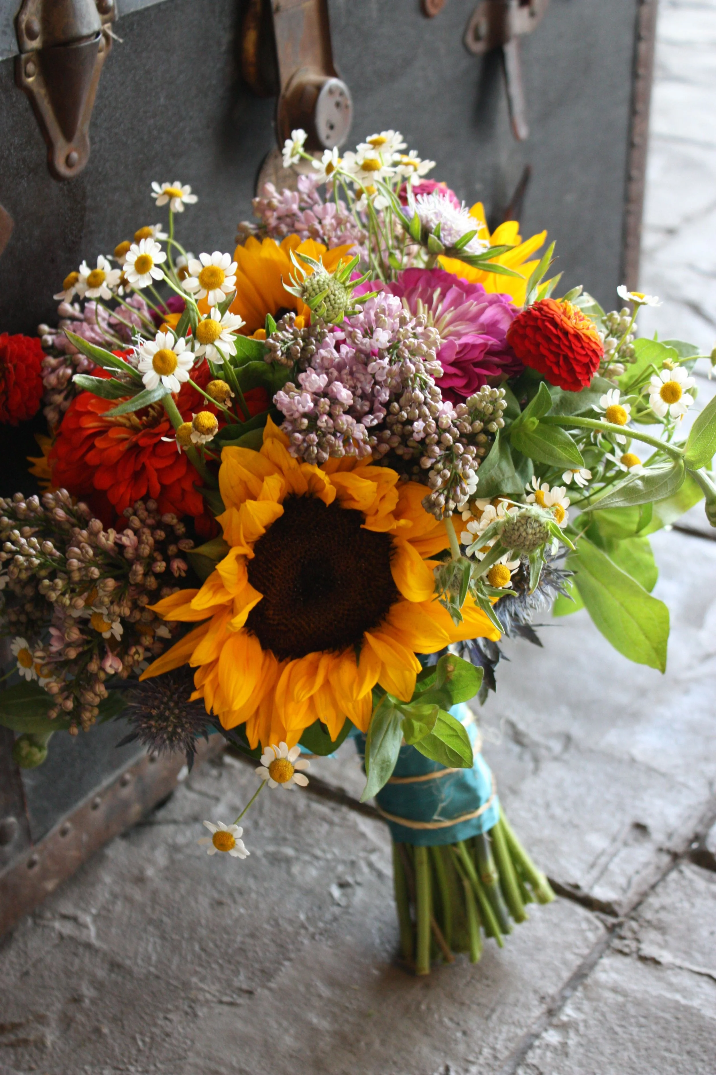 Colorful bouquet of sunflowers, daisies, zinnias, and other flowers on a gray stone floor with a dark background.