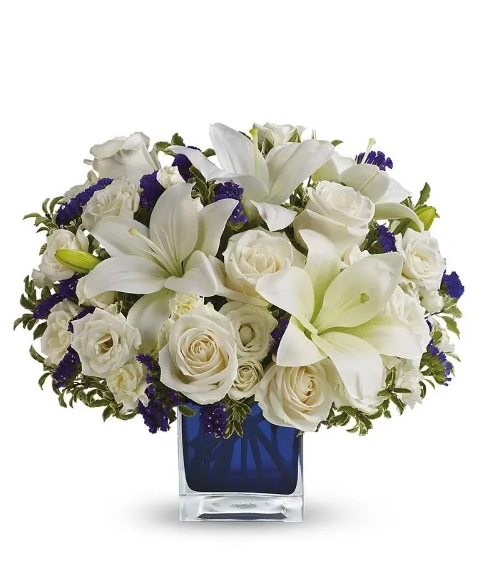 A bouquet of white lilies, roses, and purple flowers in a dark blue glass square vase.