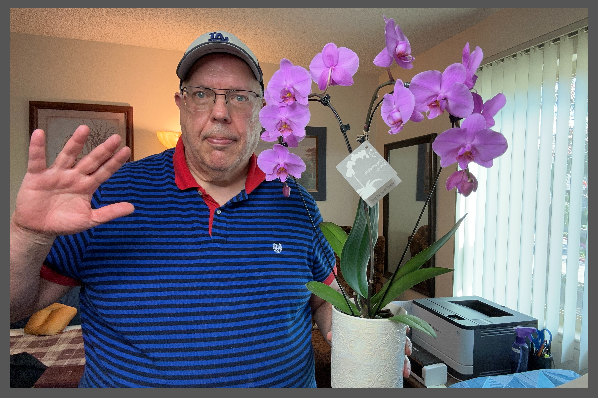 A man wearing glasses and a blue striped polo shirt with red collar, waving with his right hand, standing next to a pot of pink orchids on a table in a room with beige walls, framed artwork, and large window blinds.