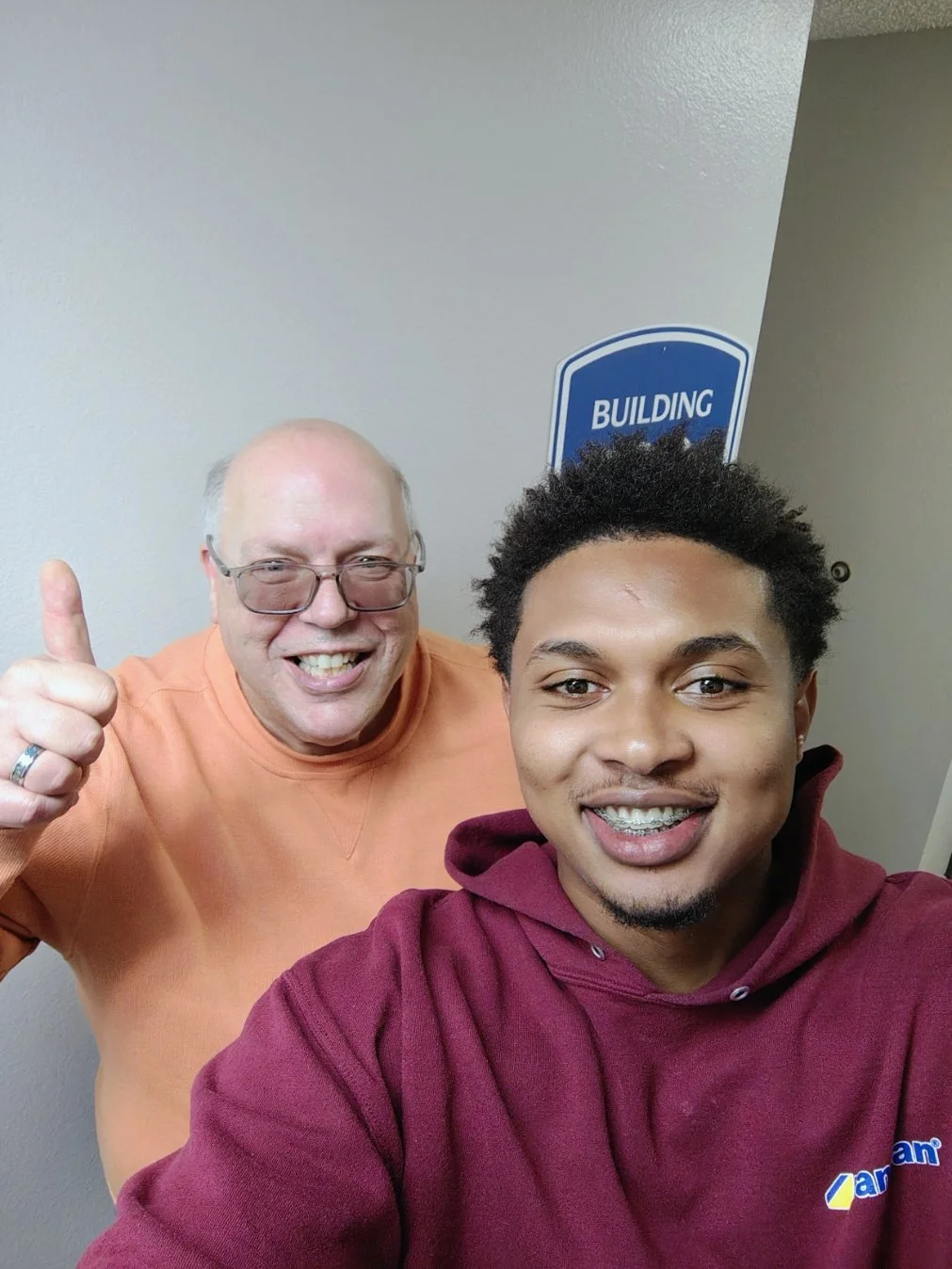 Two men taking a selfie in front of a wall with a blue sign that says 'Building'. One man is older, bald, wearing glasses and an orange shirt, giving a thumbs up. The other is younger, with curly hair, braces, and a maroon hooded sweatshirt, smiling at the camera.