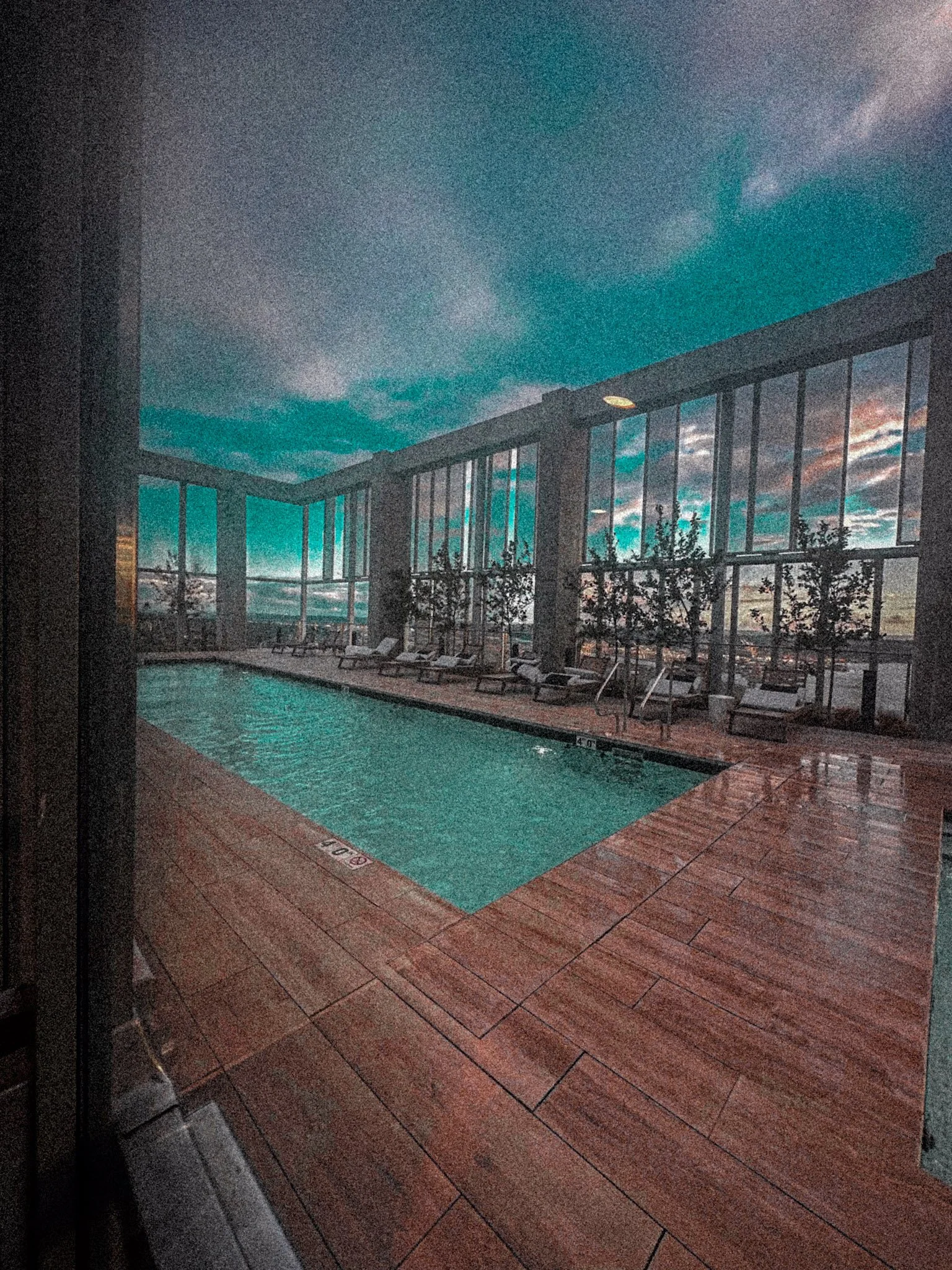 Indoor rooftop swimming pool with lounge chairs and large glass windows at sunset.