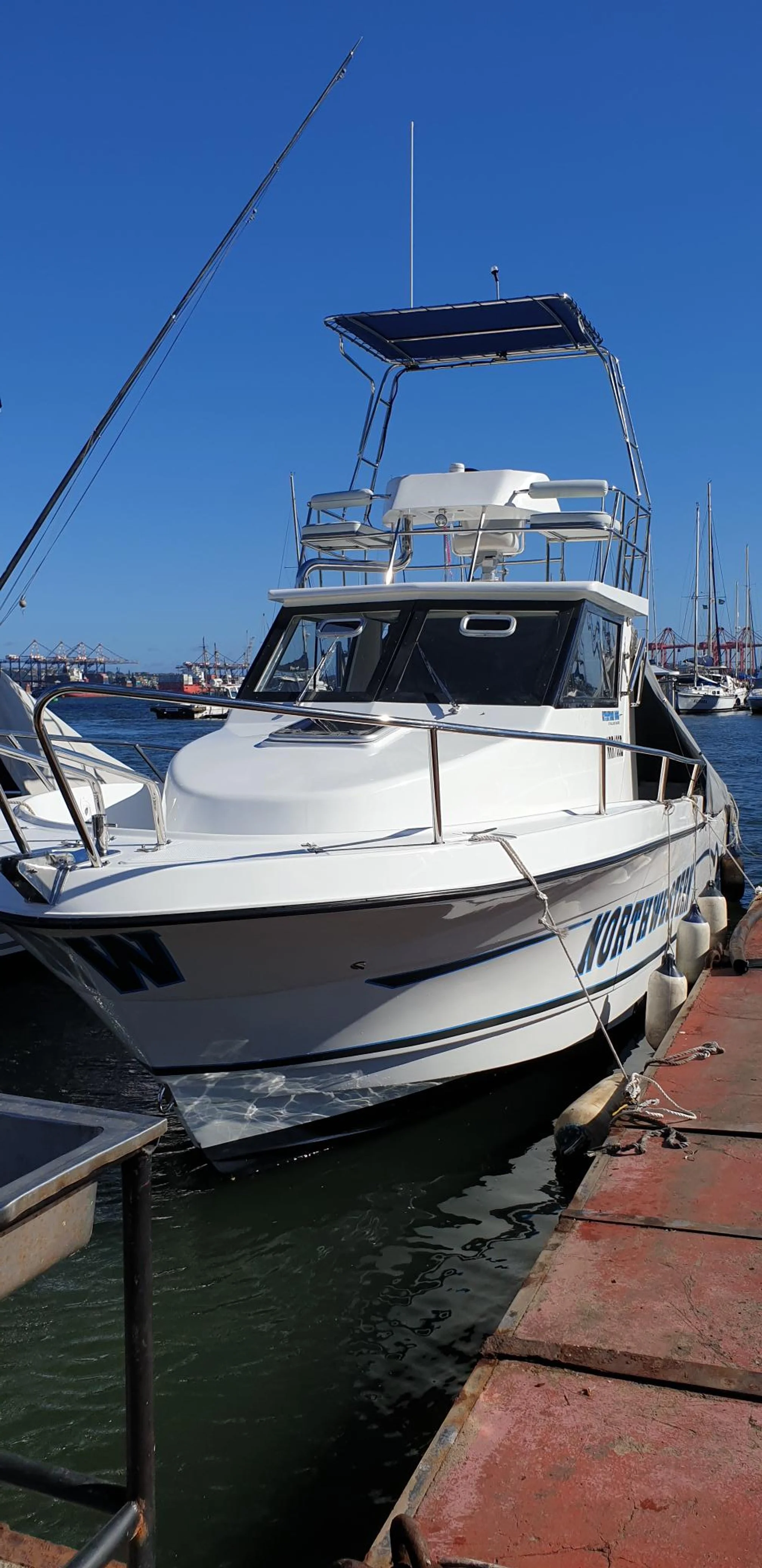 White boat docked by a pier on a sunny day with a blue sky, water, and other boats in the background.
