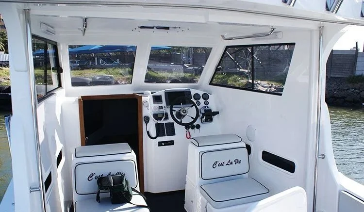 Interior of a boat with white seating, a control panel, steering wheel, and navigation equipment, docked by water.