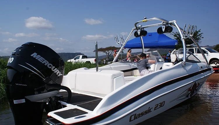 A white and black motorboat named Classic 230 with a Mercury outboard motor, docked on a body of water with a group of people on board. The boat has a blue canopy and wakeboard racks, with parked cars and a grassy area in the background.