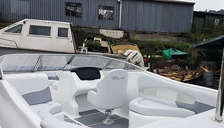 A small white boat with seating and a captain's chair, parked outdoors near a warehouse and old boats in the background.