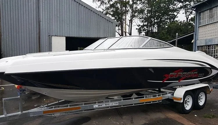 A white and black speedboat on a trailer parked outside near industrial buildings and trees.