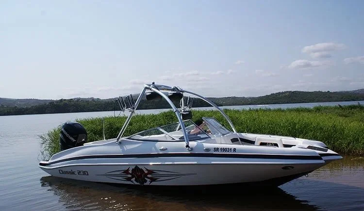 A white motorboat named Classic 230 anchored near the shoreline in a lake, with grassy banks and distant hills under a partly cloudy sky.