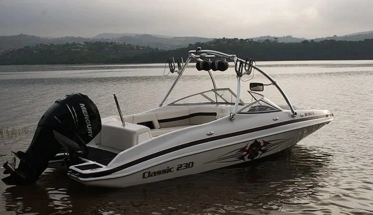 A small white motorboat with black and red accents floats on calm water, with a Mercury outboard engine at the stern, and a metal wakeboard tower above the boat. The background shows distant hills under an overcast sky.