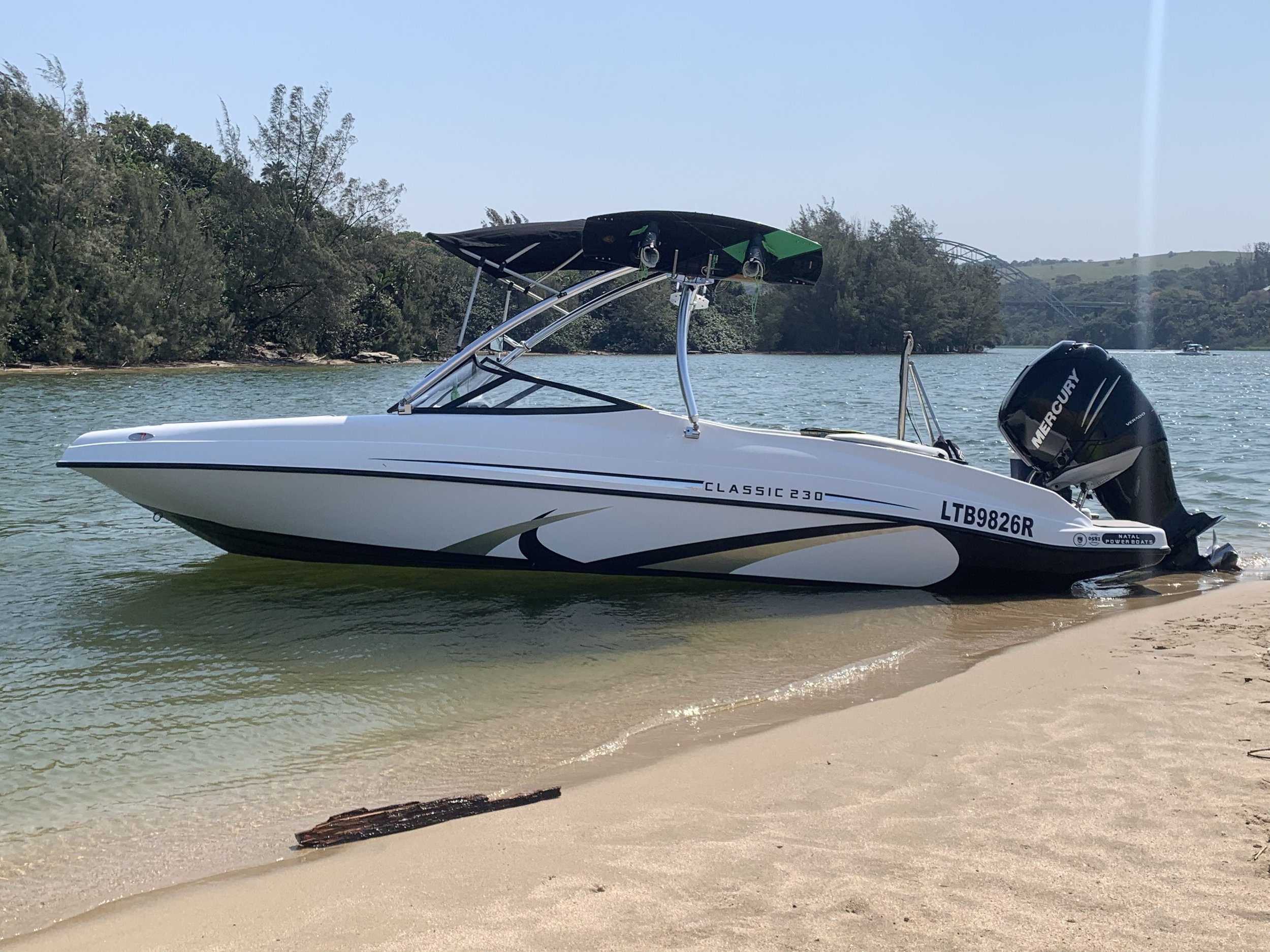 A white speedboat with a Mercury outboard motor, partially on the sandy shore of a lake, with trees and a bridge in the background.