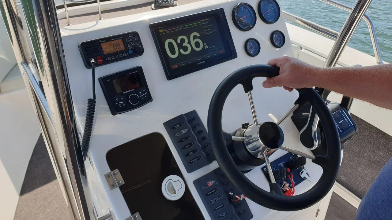 View of a boat's control dashboard with a steering wheel, digital and analog gauges, and electronic devices, on open water.