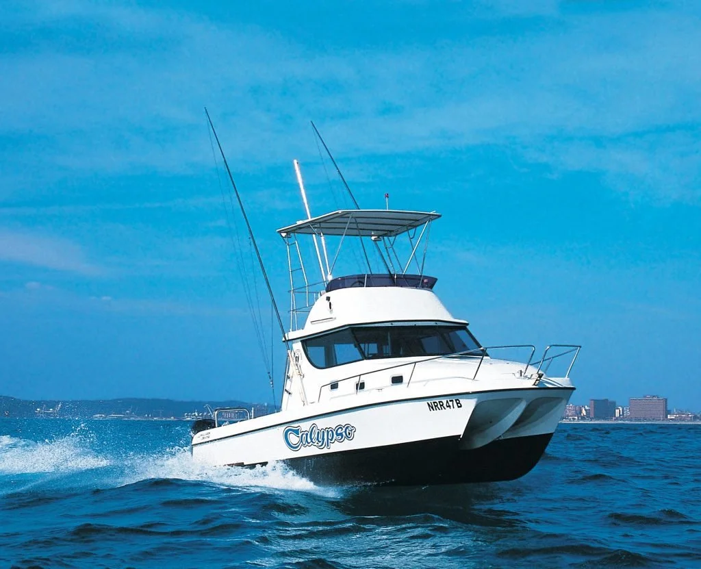 White motor yacht named Calypso sailing on blue water under a partly cloudy sky with a cityscape in the background.