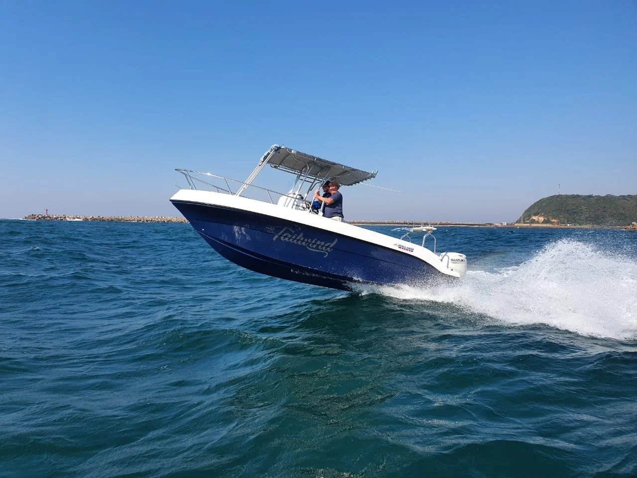 A person driving a white and blue boat on the water during a sunny day, with a landmass in the background.