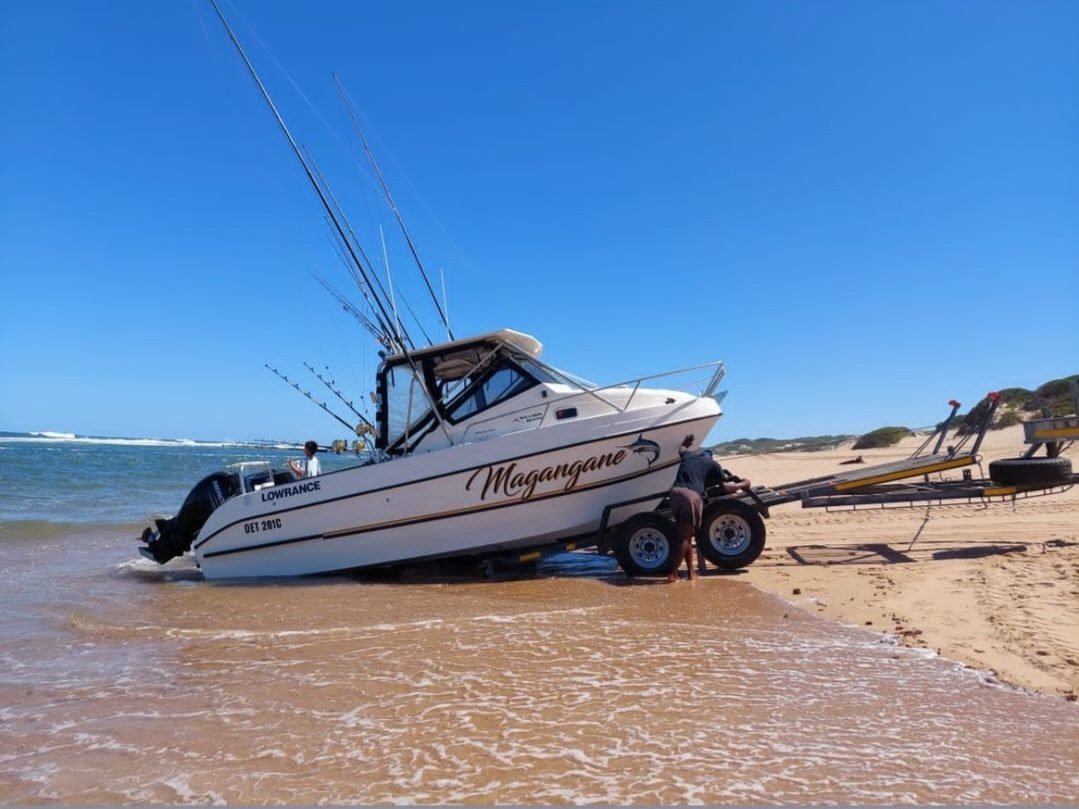 A boat named Magangane being launched into the ocean on a sandy beach with a trailer attached, under a clear blue sky.