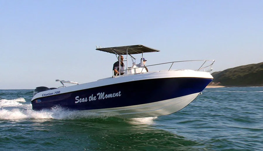 A boat named 'Seas the Moment' is speeding on the water with two people on board, near a hilly coastline.